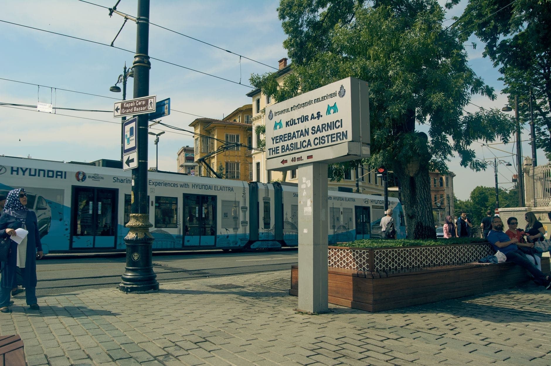 Pemandangan jalan siang hari di Istanbul dengan tram, tanda yang jelas menuju atraksi utama, dan orang-orang yang menunggu di dekat bangku di bawah pohon.