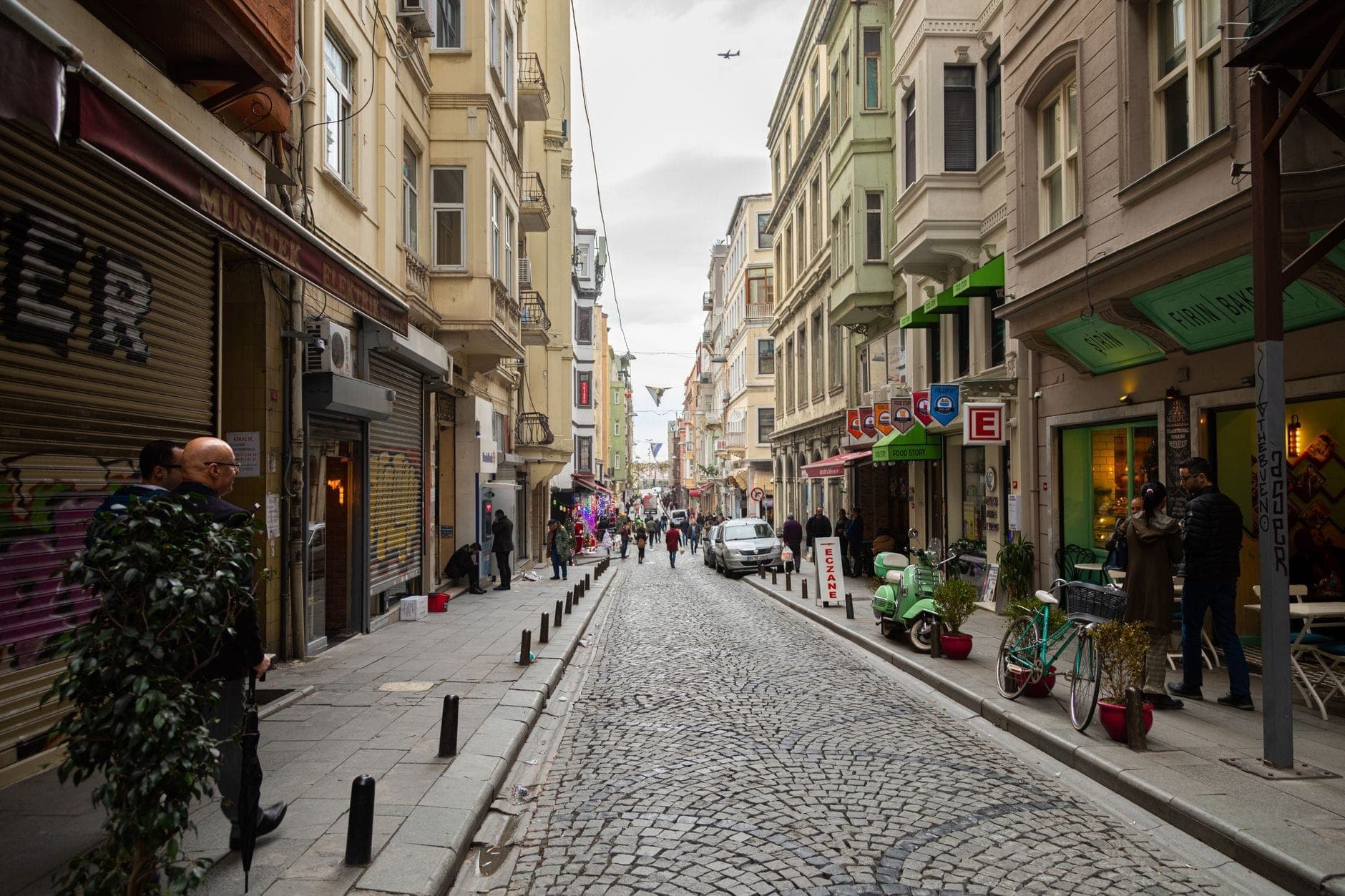 Pemandangan jalanan khas Istanbul dengan jalan berbatu, orang-orang berjalan, toko-toko lokal, dan apartemen, yang menggambarkan kawasan perkotaan.