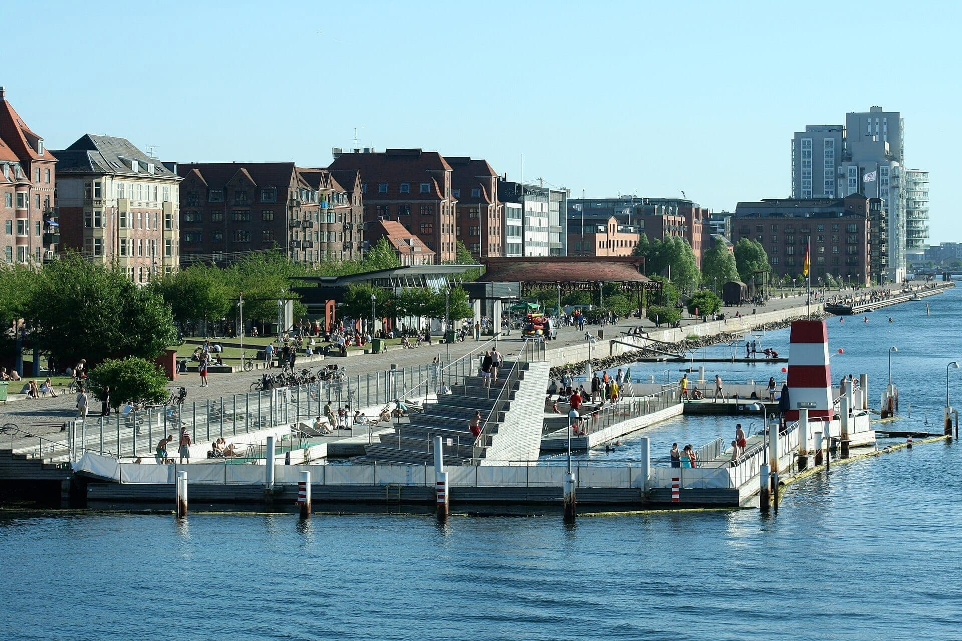 Blick auf das Islands Brygge Harbour Bath in Kopenhagen mit Menschen, die an einem sonnigen Tag die Pools, den Fluss und die angrenzenden Stadtgebäude genießen.