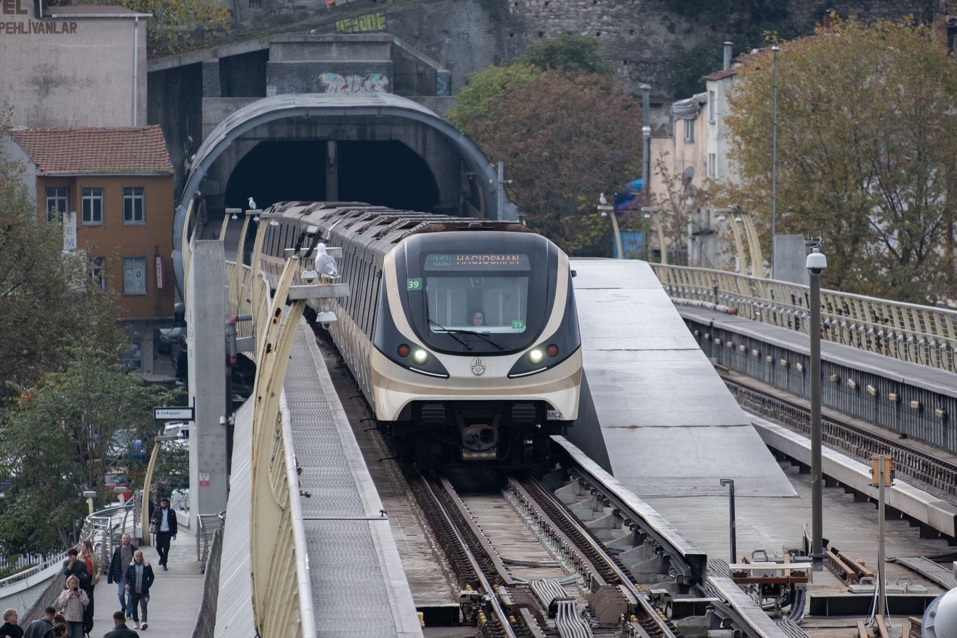 Treno della Metro di Istanbul che attraversa un ponte verso un tunnel con edifici della città sullo sfondo e persone che camminano nelle vicinanze.