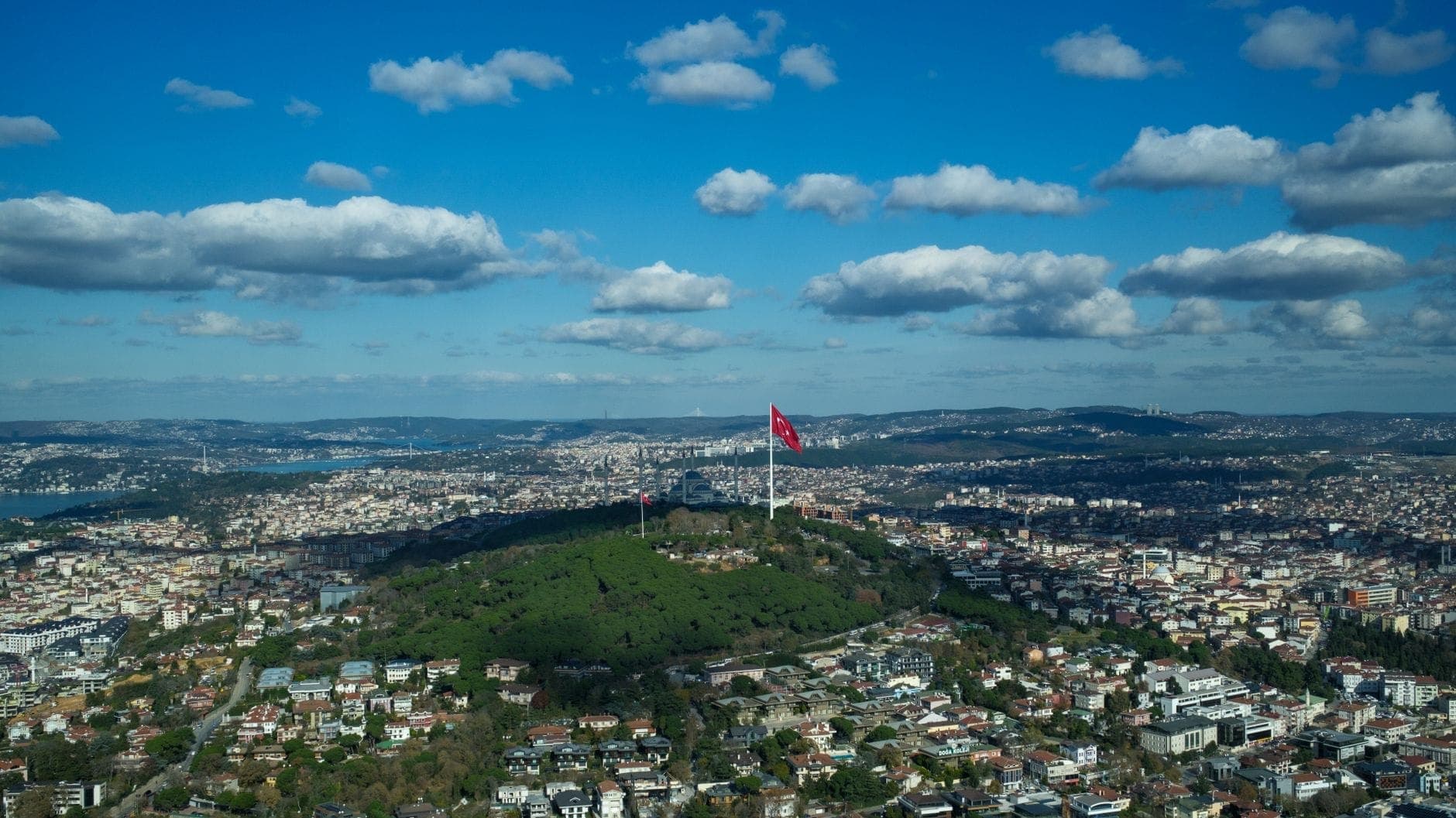Luftaufnahme der asiatischen Seite von Istanbul mit dem Çamlıca Hill, türkischen Flaggen, Grünflächen und einer weitläufigen Stadtlandschaft unter blauem Himmel mit Wolken.