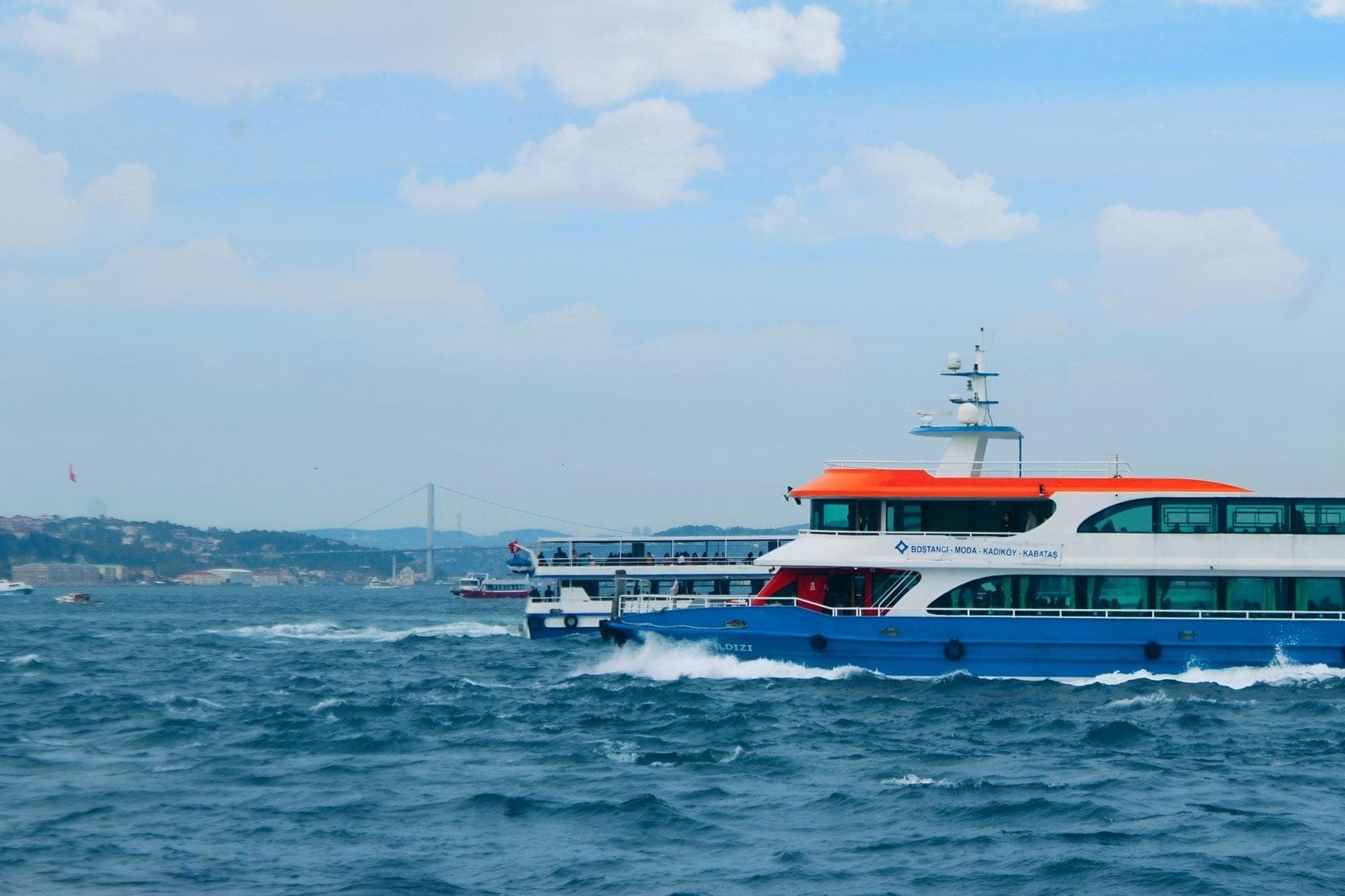 Eine Stadtfähre überquert den Bosporus in Istanbul mit der Bosporus-Brücke und der Skyline der Stadt im Hintergrund.