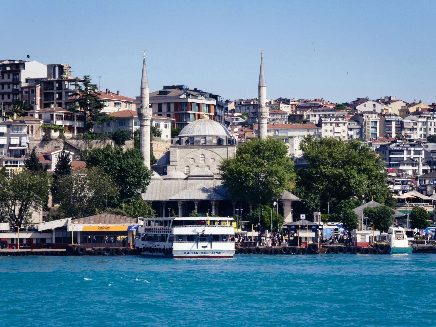 Uferpromenade von Üsküdar mit einer historischen Moschee, Fährschiffen und Wohngebäuden im Hintergrund an einem sonnigen Tag.