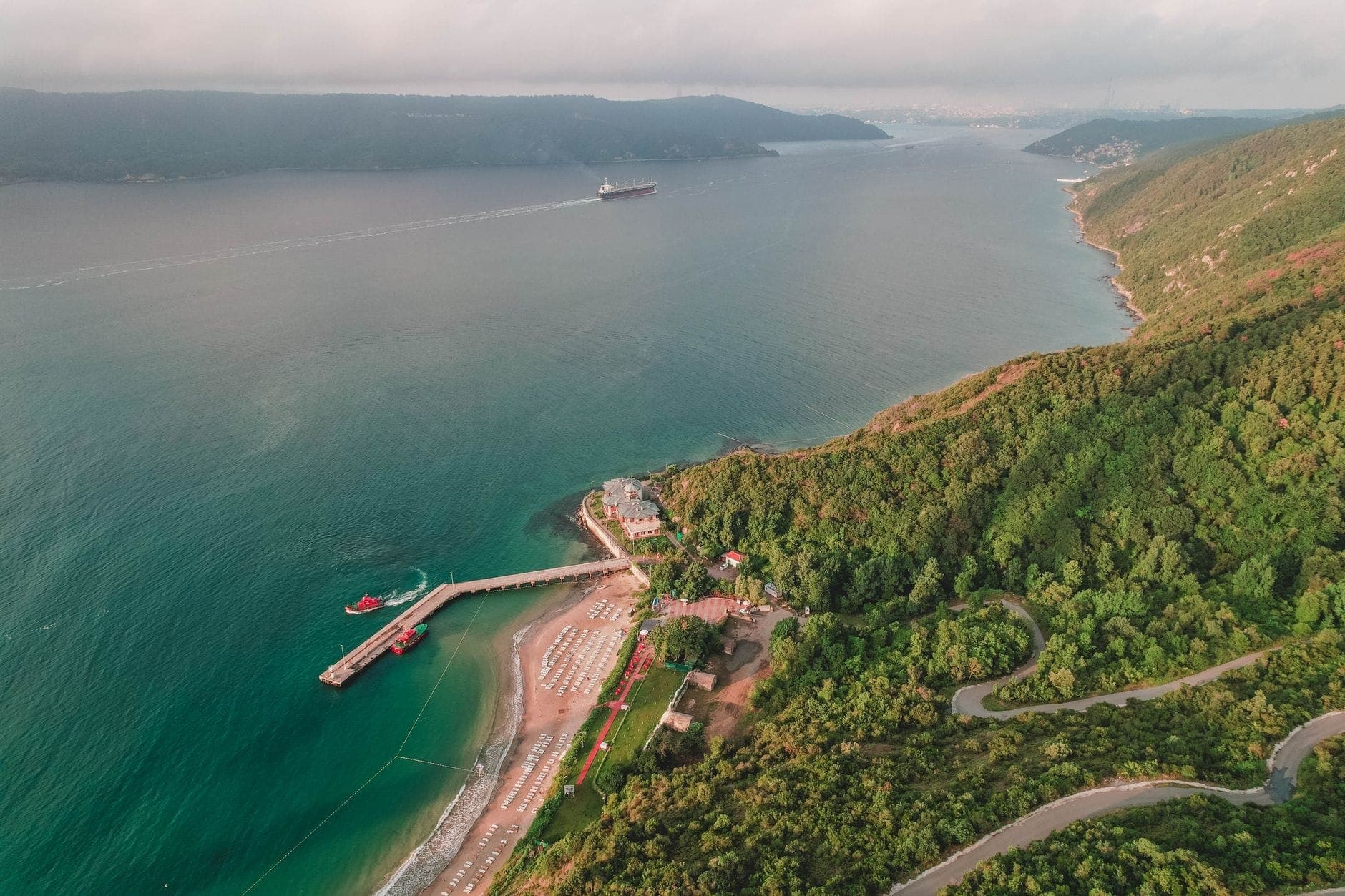 Luftaufnahme einer malerischen Küste bei Istanbul mit grünen Hügeln, einem Sandstrand, einem Pier und einem Frachtschiff in der Ferne auf ruhigem blauem Wasser.