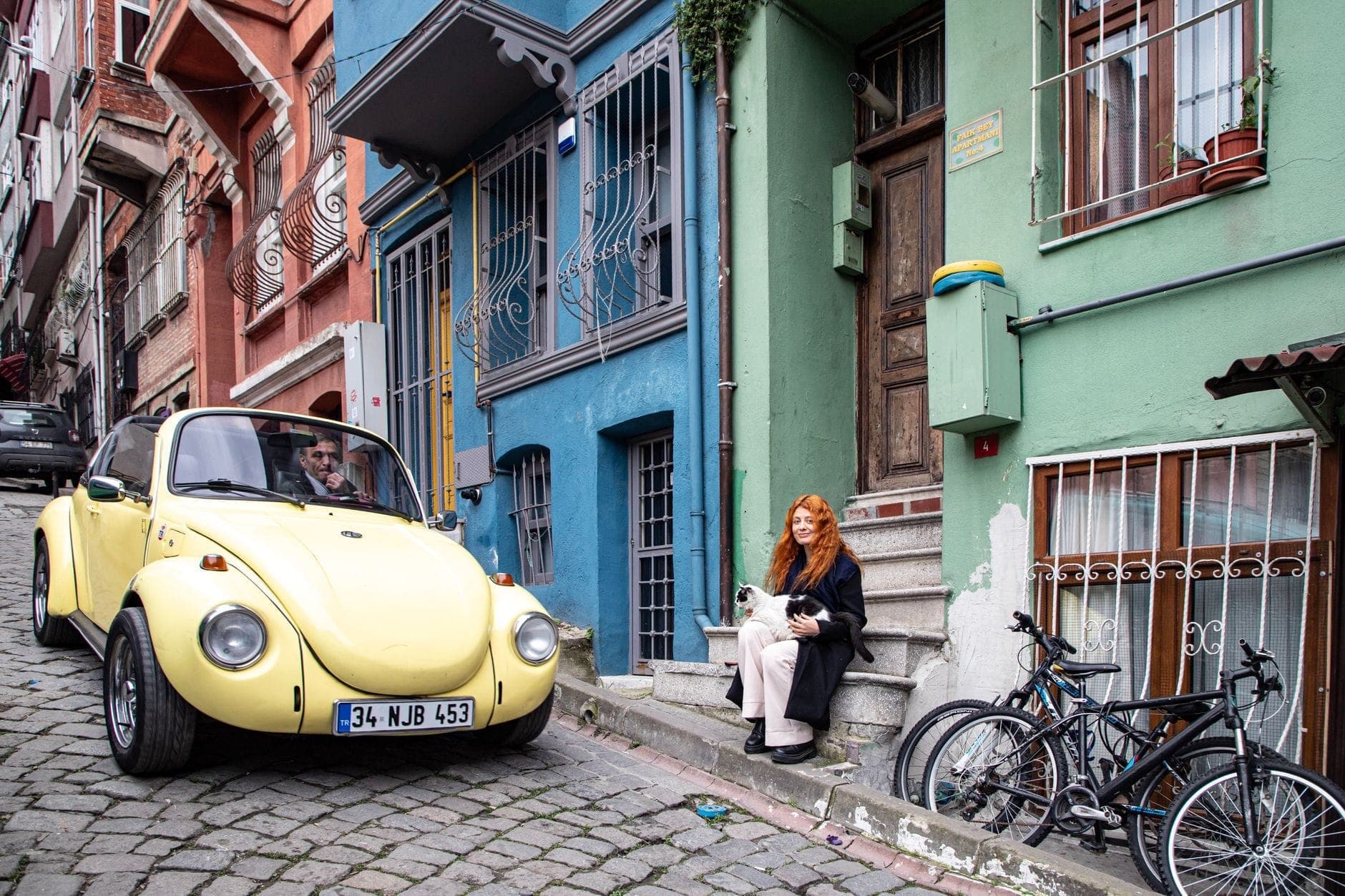 Frau, die auf einer Treppe in einer bunten Straße in Istanbul sitzt und eine Katze streichelt, mit historischen Gebäuden und einem gelben Auto in der Nähe.