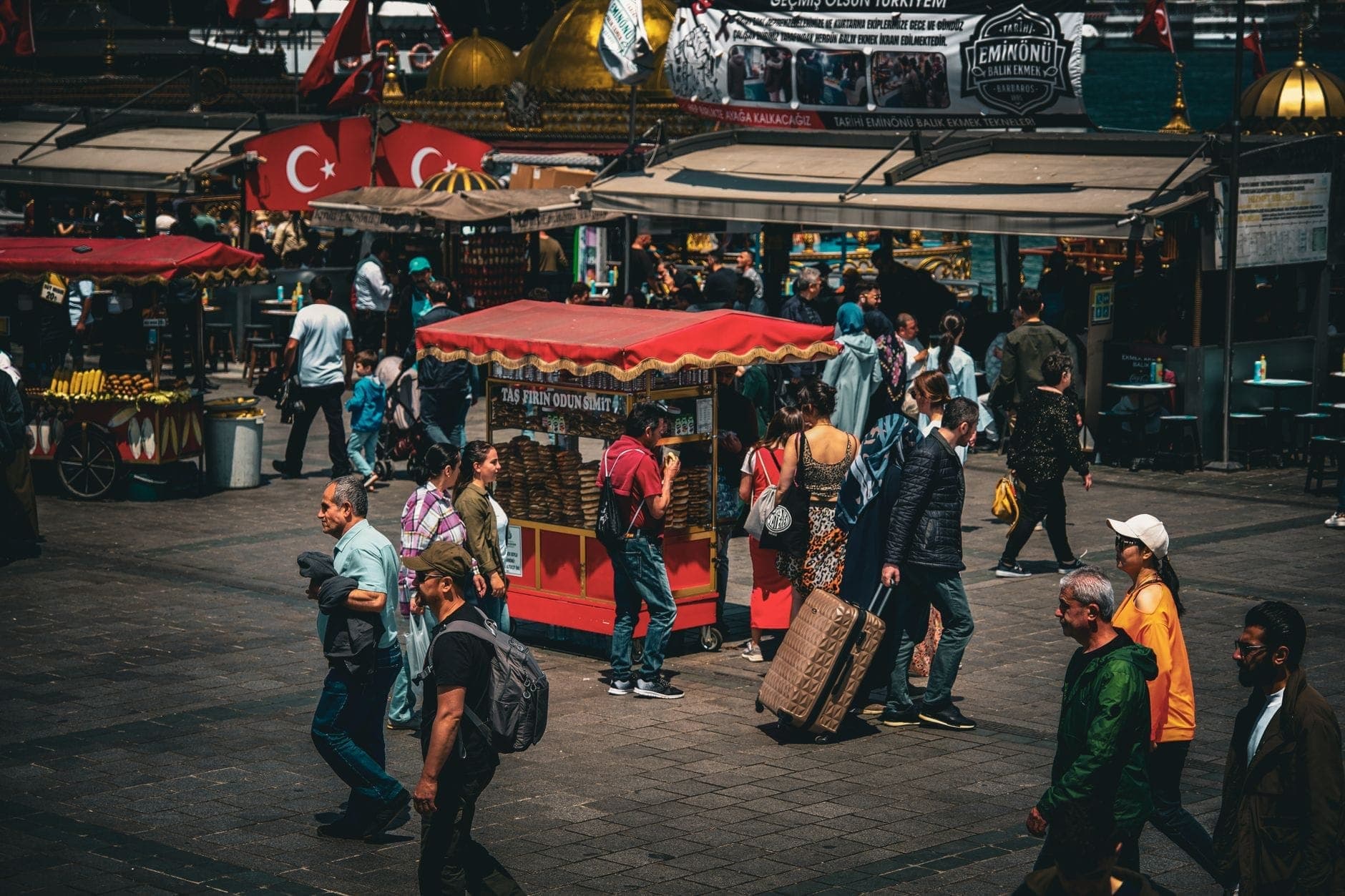 Pasar jalanan Istanbul yang ramai dengan kios makanan Turki, gerobak merah, bendera Turki, dan orang-orang berjalan, menangkap kehidupan kuliner jalanan.