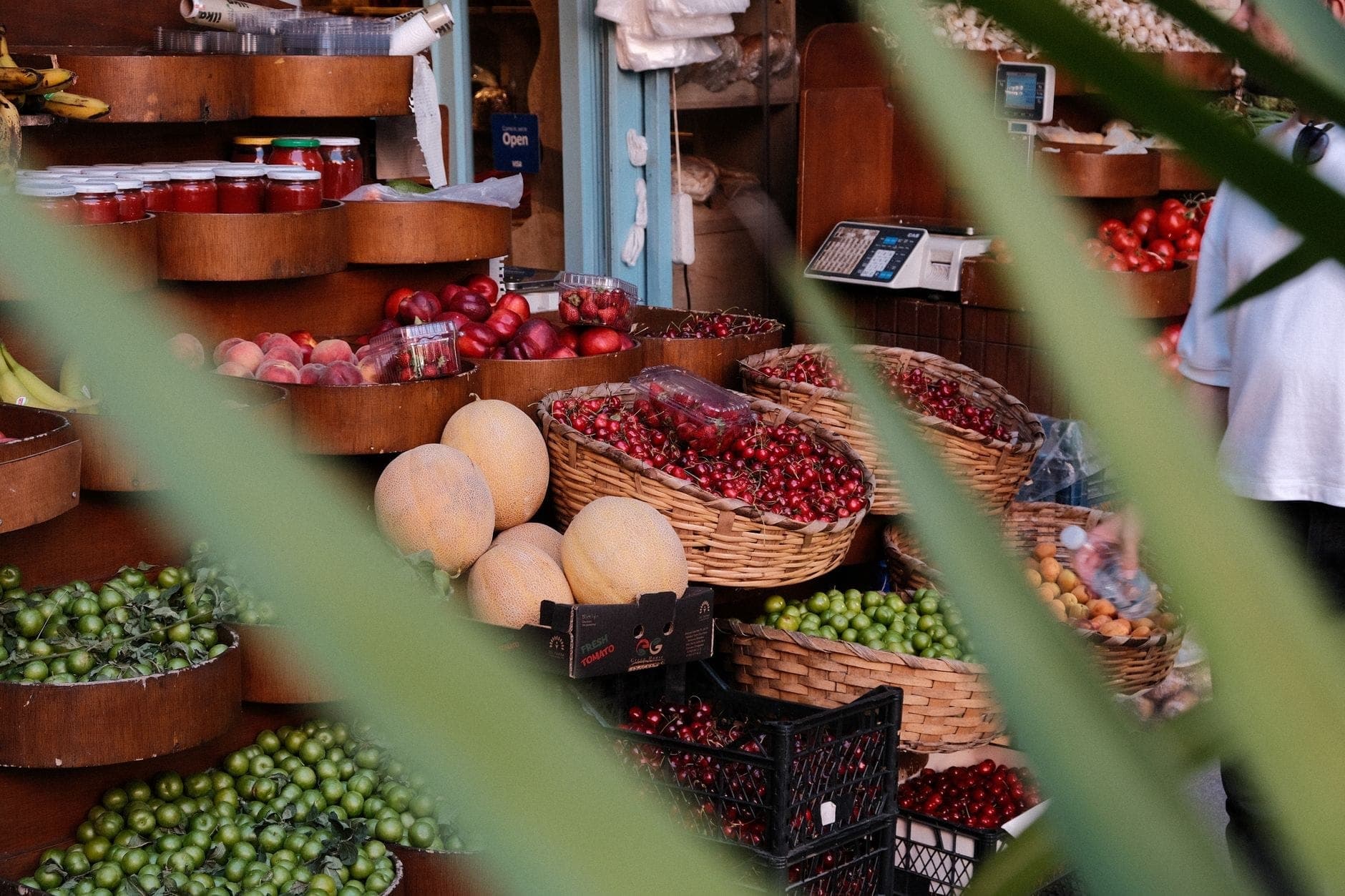 Tampilan close-up kios pasar makanan yang semarak di Istanbul yang dipenuhi buah-buahan segar, sayuran, dan toples, dilihat melalui tanaman berdaun.
