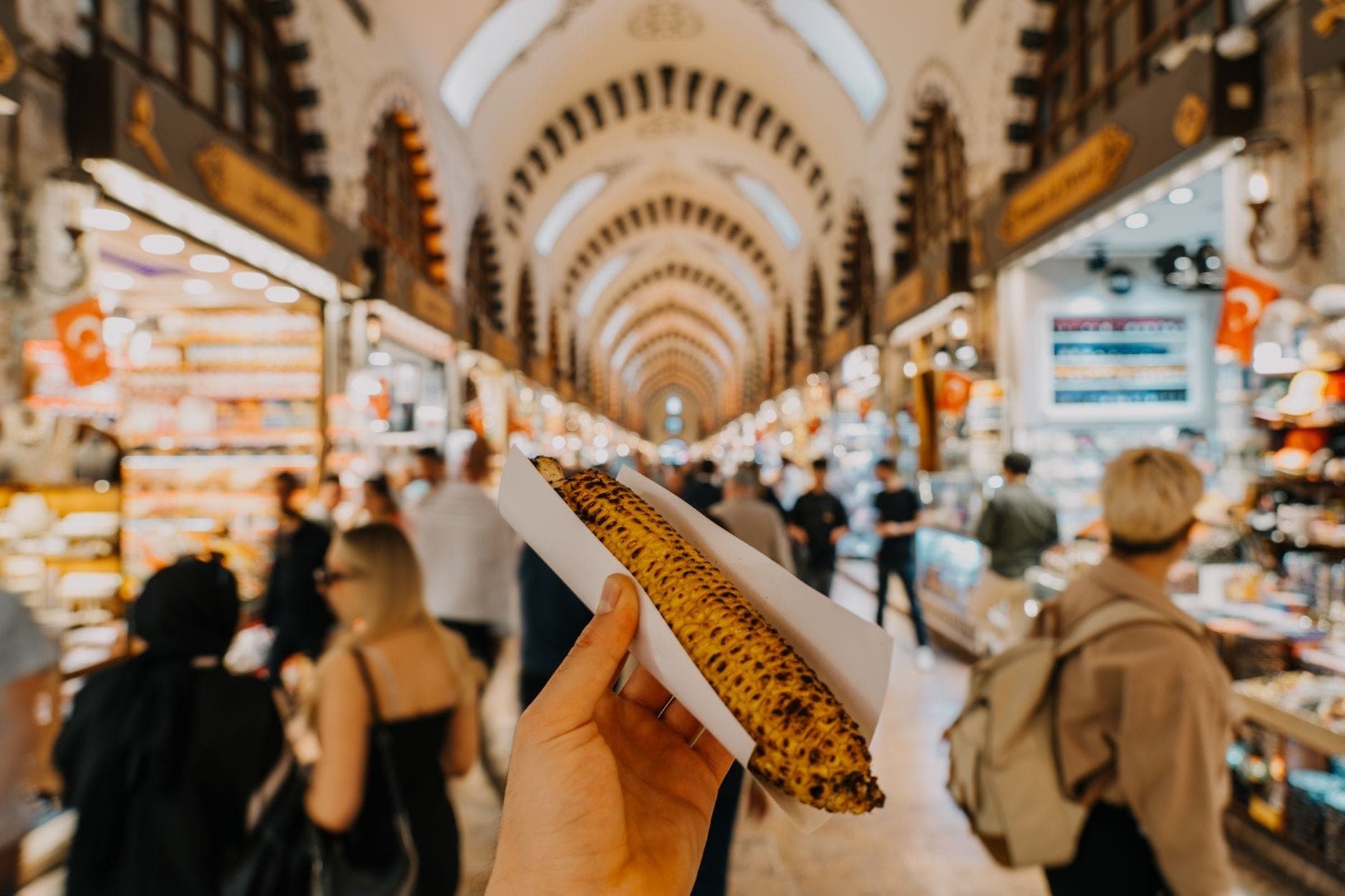 Seseorang memegang jagung bakar di lorong-lorong ramai bazaar rempah-rempah dalam ruangan yang terkenal di Istanbul, dengan para pembeli di latar belakang.