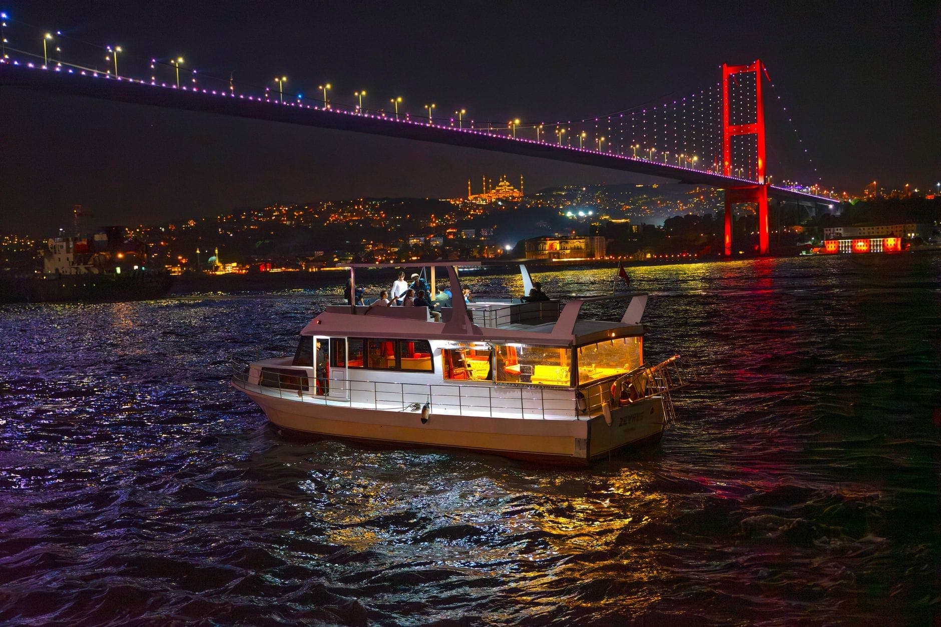 Ein hell erleuchtetes Boot, das nachts auf dem Bosporus fährt, mit der beleuchteten Bosporus-Brücke und der Skyline von Istanbul im Hintergrund.