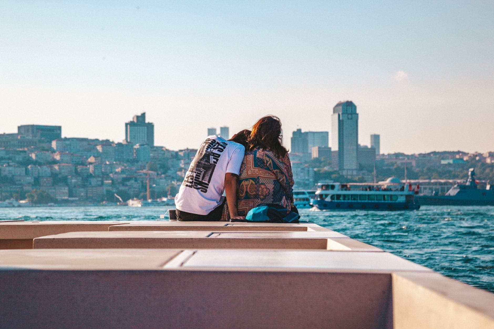Ein Paar sitzt eng beieinander auf einer Bank am Wasser in Istanbul und blickt auf die Skyline der Stadt und die Boote auf dem Bosporus unter einem strahlend blauen Himmel.