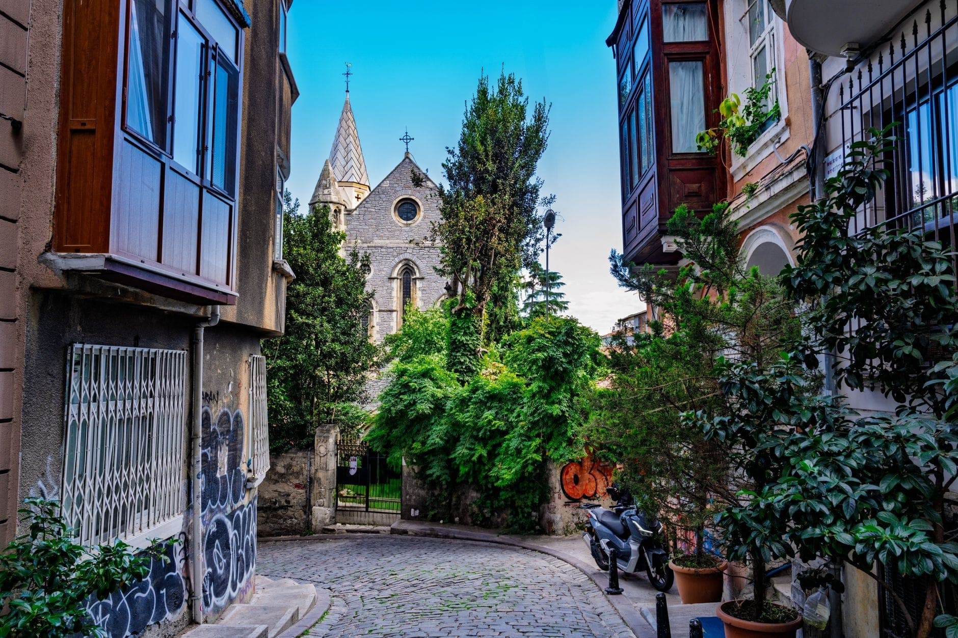 Eine ruhige, enge Kopfsteinpflasterstraße in Istanbul, gesäumt von alten Häusern und Grün, die zu einer versteckten Steinkirche mit spitzen Türmen und einem Kreuz führt – in strahlendem Tageslicht.