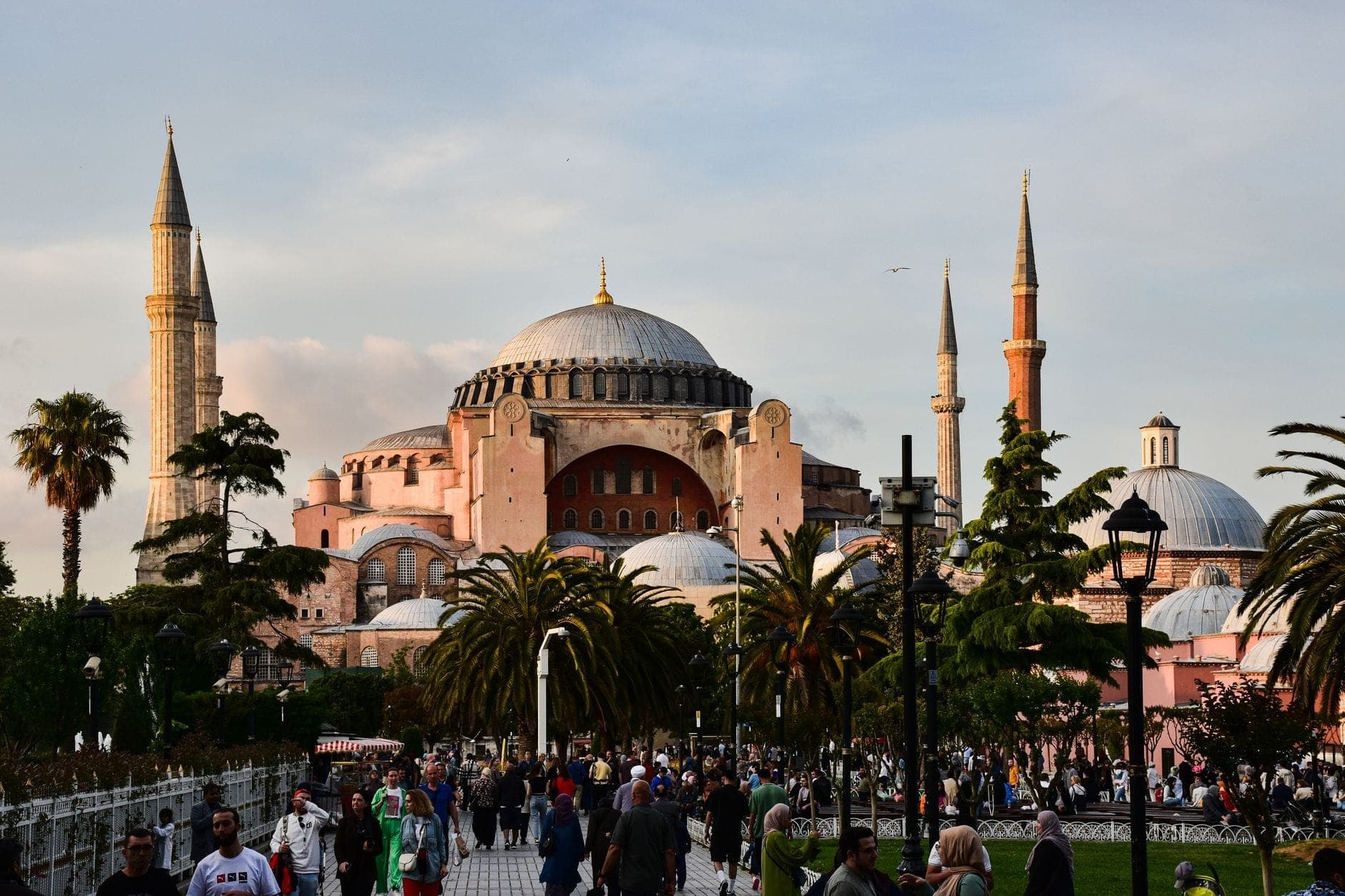 Multitudes de personas caminan y socializan cerca de la Hagia Sophia en Estambul, con vegetación y el histórico edificio con cúpula al fondo.