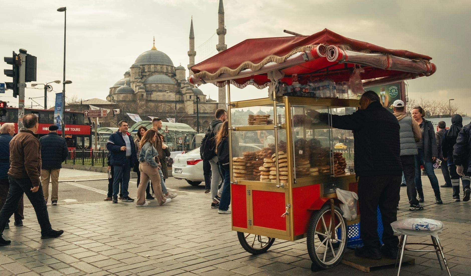 Escena callejera con un carrito de simit, personas caminando y una prominente mezquita con cúpulas y minaretes al fondo, probablemente junto al Golden Horn.