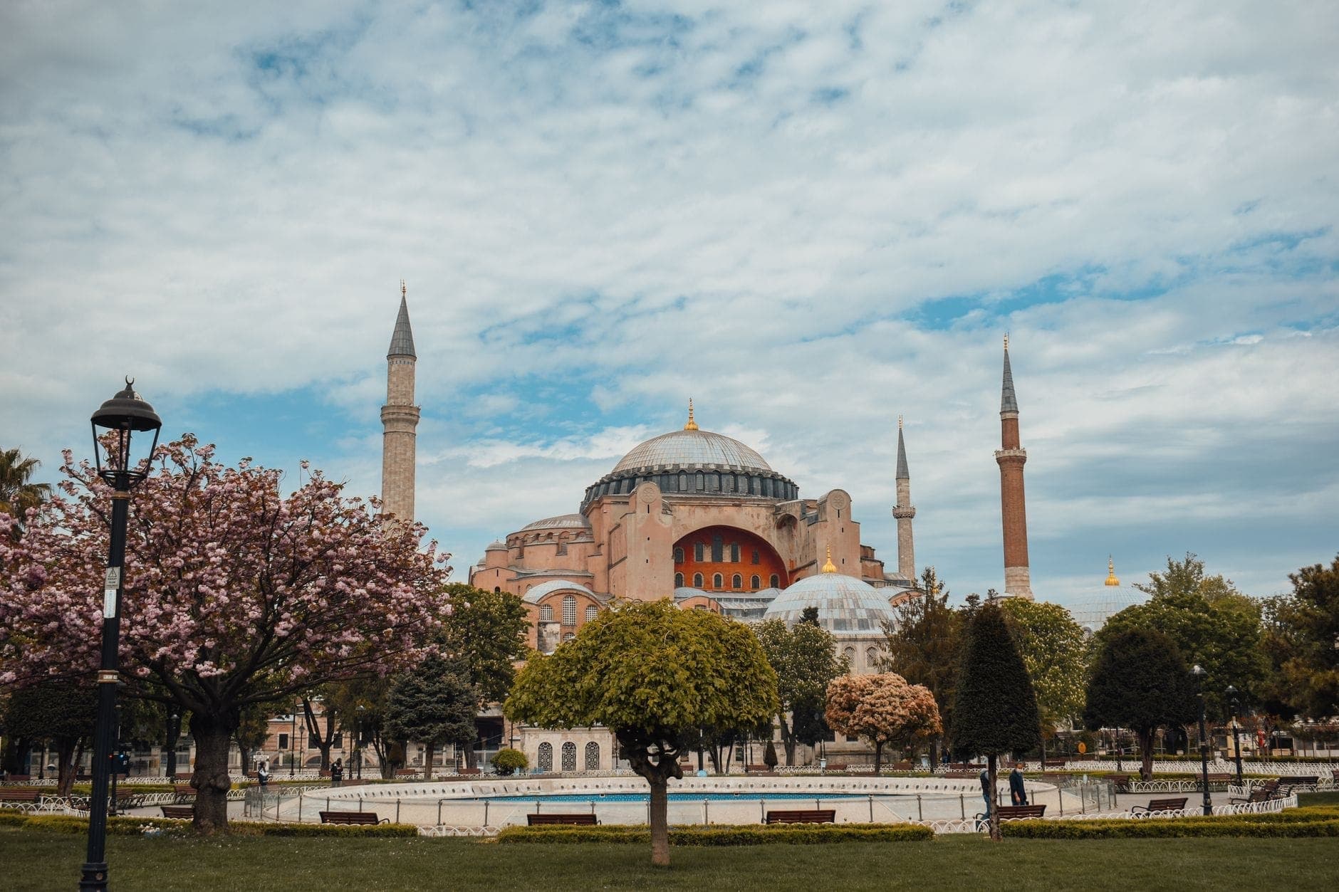 Hagia Sophia with blooming spring trees and people strolling in the foreground on a cloudy day in Istanbul.