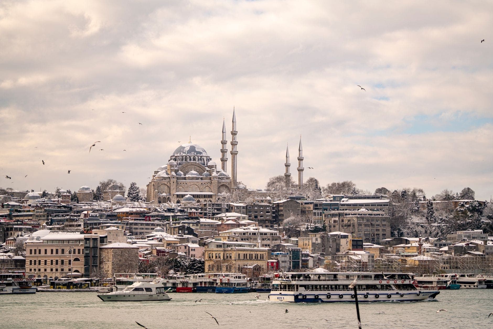 Panoramablick auf Istanbul im Winter mit historischen Moscheen, schneebedeckten Dächern, Bosporus-Fähren und Möwen unter einem dramatischen Winterhimmel.