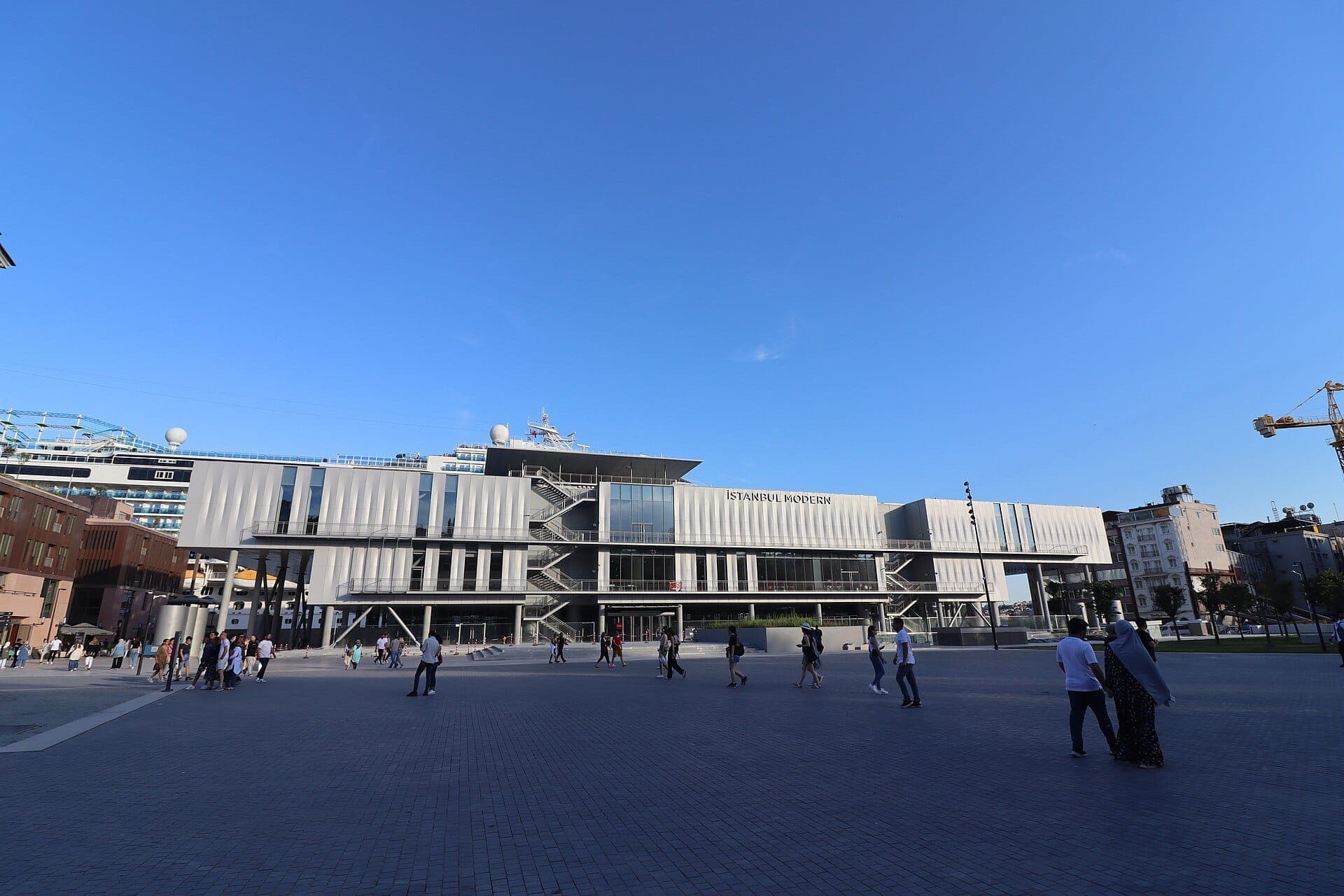 Weitwinkelansicht der eleganten, modernen Fassade des Museums Istanbul Modern mit Menschen, die bei klarem blauem Himmel über den weitläufigen Platz spazieren.