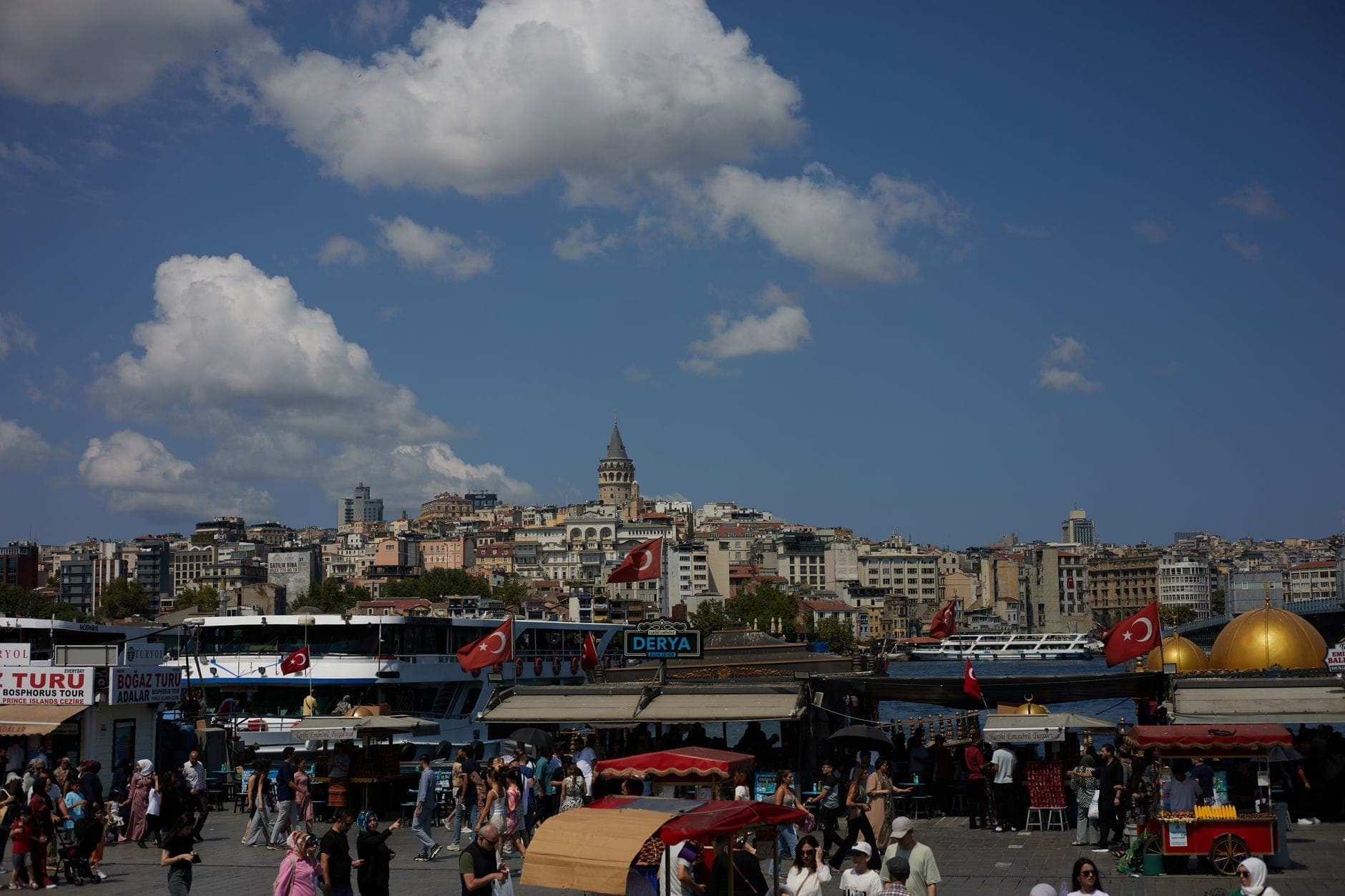 Alun-alun luar ruangan yang ramai di Istanbul dengan kerumunan wisatawan dan warga lokal, banyak kios pedagang, serta Menara Galata yang terlihat di latar belakang.