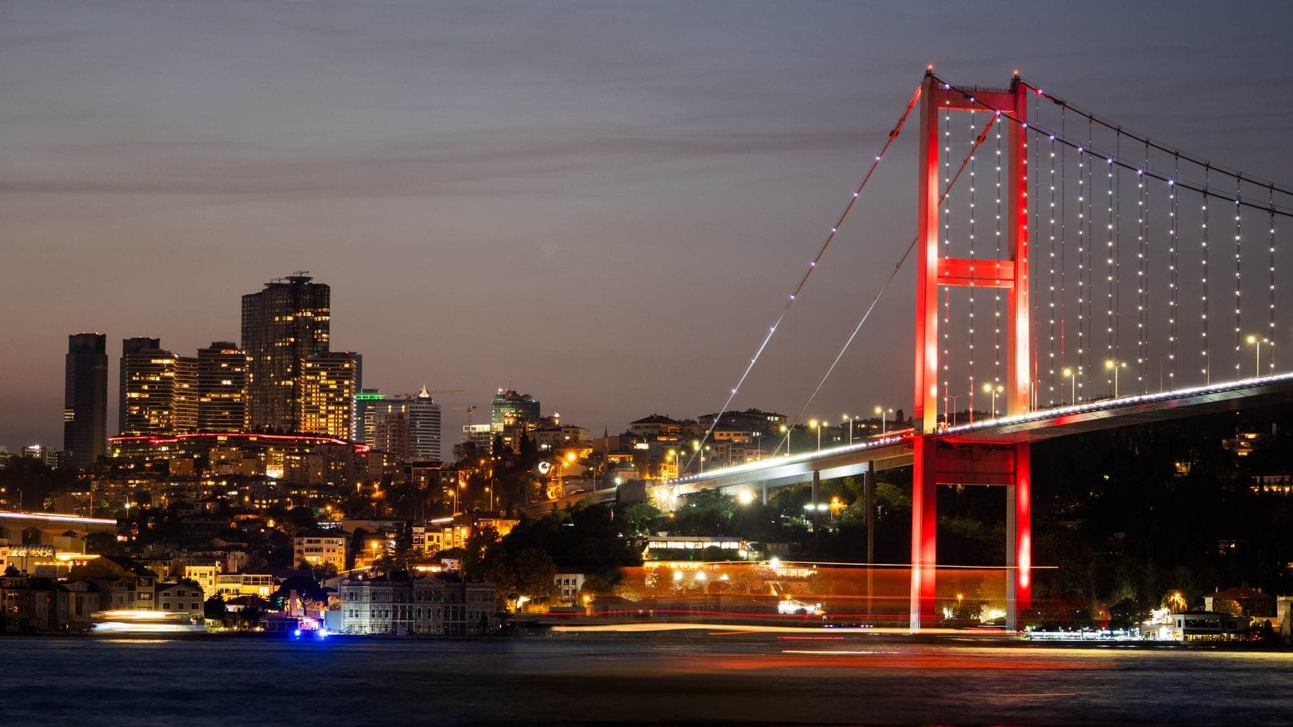 Vista nocturna del Puente del Bósforo y las luces de la ciudad junto al agua, con edificios de lujo y una animada escena ribereña.