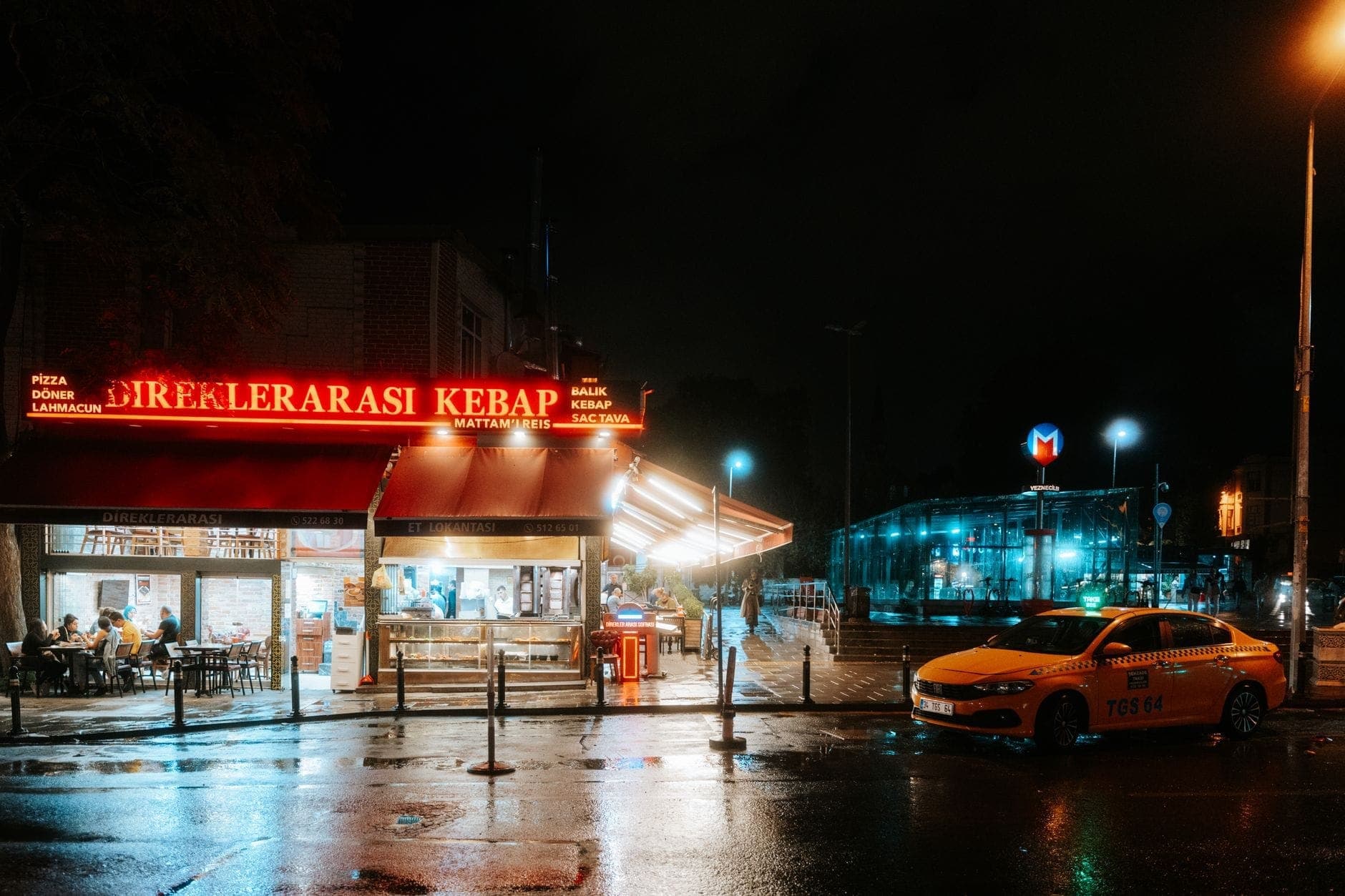 Un restaurante de kebab con un letrero de neón brillante y un taxi amarillo fuera en una noche lluviosa en Estambul, con la entrada al metro visible.