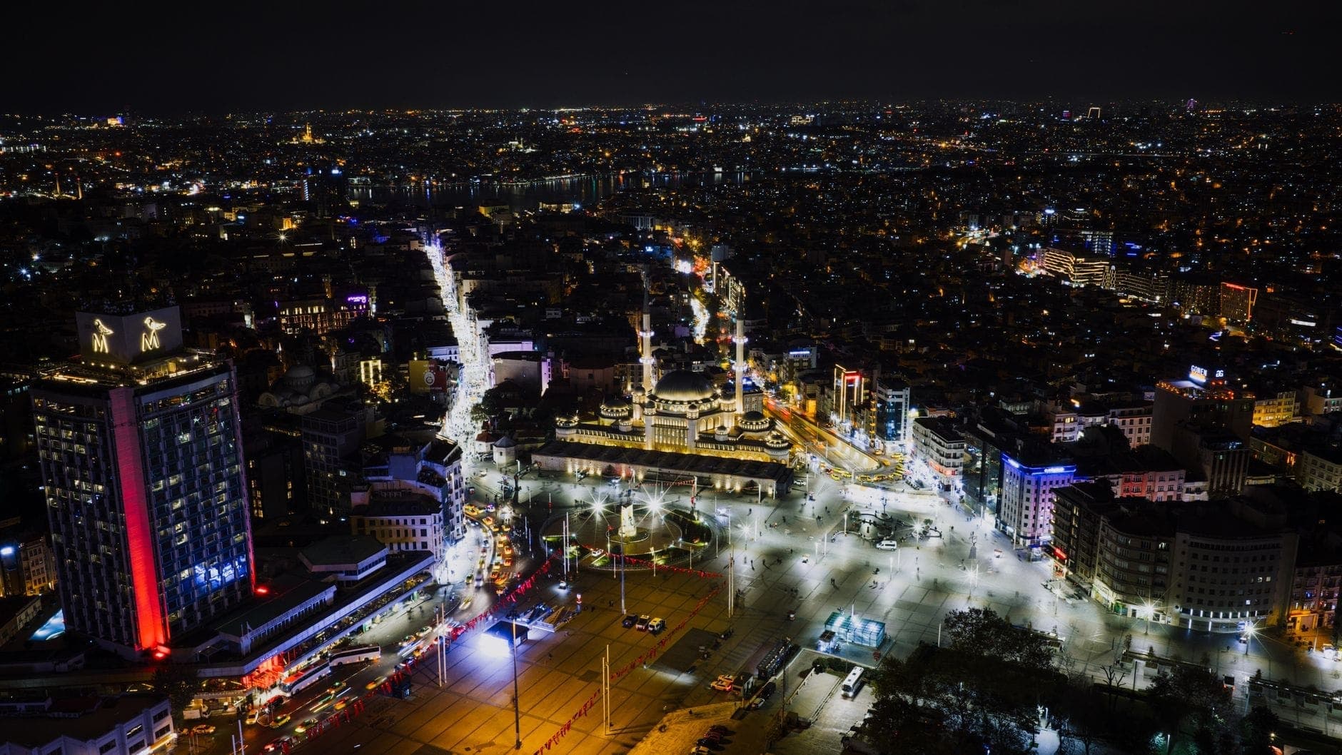 Nächtliche Luftaufnahme von Istanbuls Stadtzentrum mit hell erleuchteten Straßen, lebhaften Gebäuden und dem belebten Taksim-Platz vor einer weit ausgedehnten Stadtlandschaft.