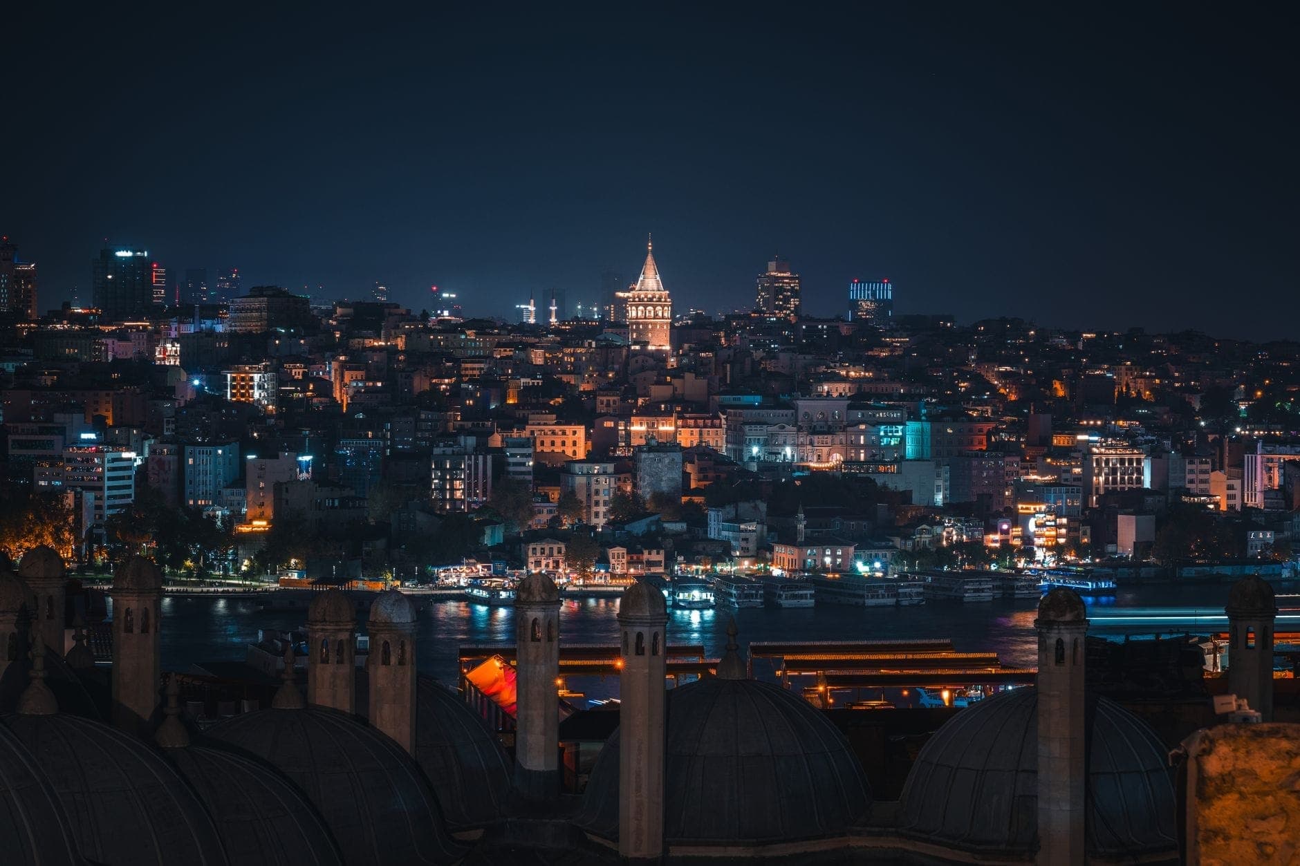 Vista nocturna de Estambul con la iluminada Torre Gálata, las luces de la ciudad y cúpulas de tejados en primer plano.