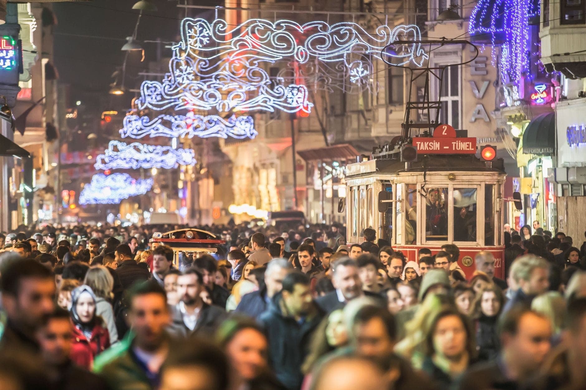 La concurrida avenida Istiklal de noche con luces festivas, un tranvía nostálgico rojo y multitudes de personas disfrutando del animado ambiente nocturno de Estambul.