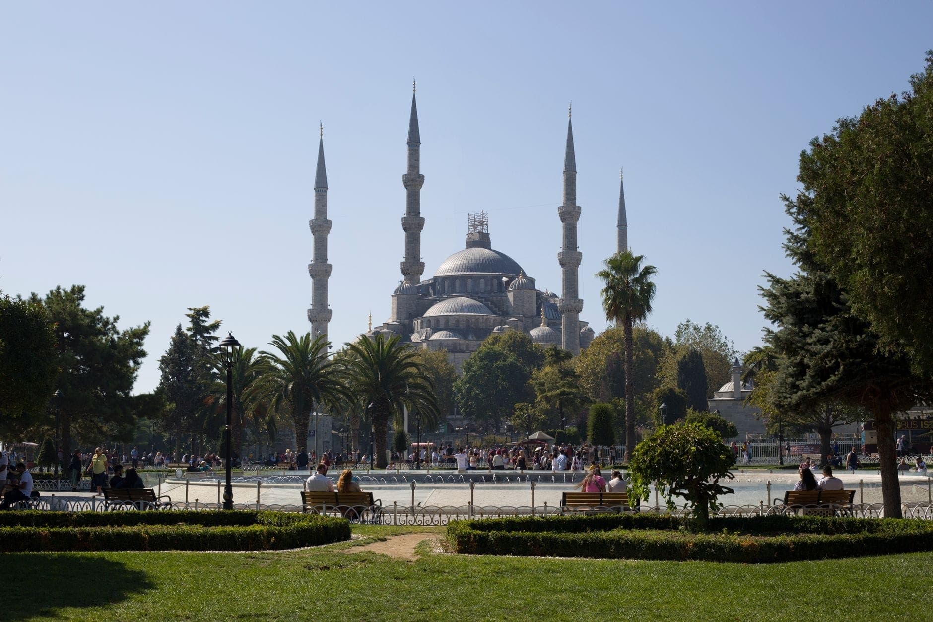 Vista amplia de la Mezquita Azul de Estambul con seis minaretes y jardines en frente, personas descansando en bancos del parque en primer plano, día soleado.