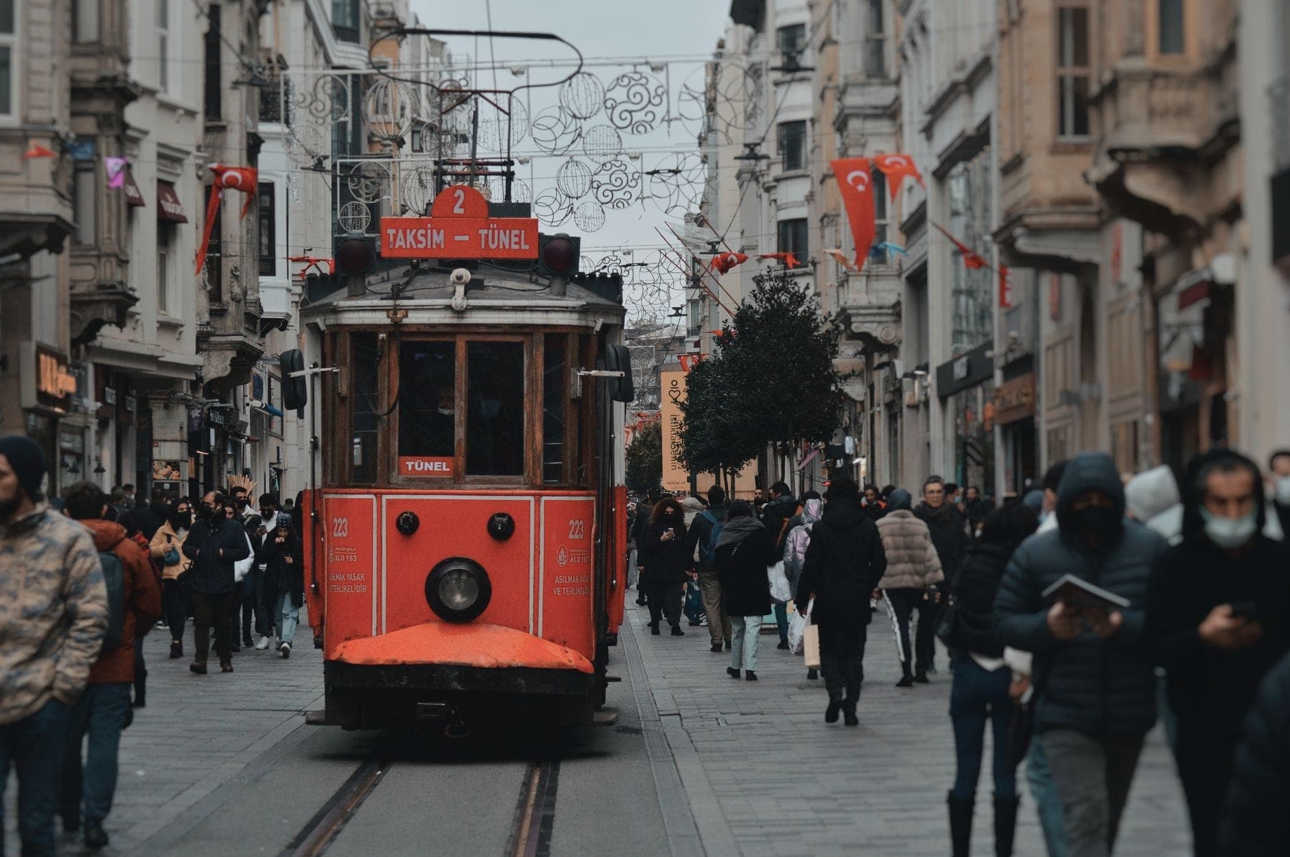 Культовый красный трамвай Taksim-Tünel едет по оживлённому проспекту İstiklal в районе Beyoğlu, Стамбул, в окружении пешеходов и исторических зданий XIX века.