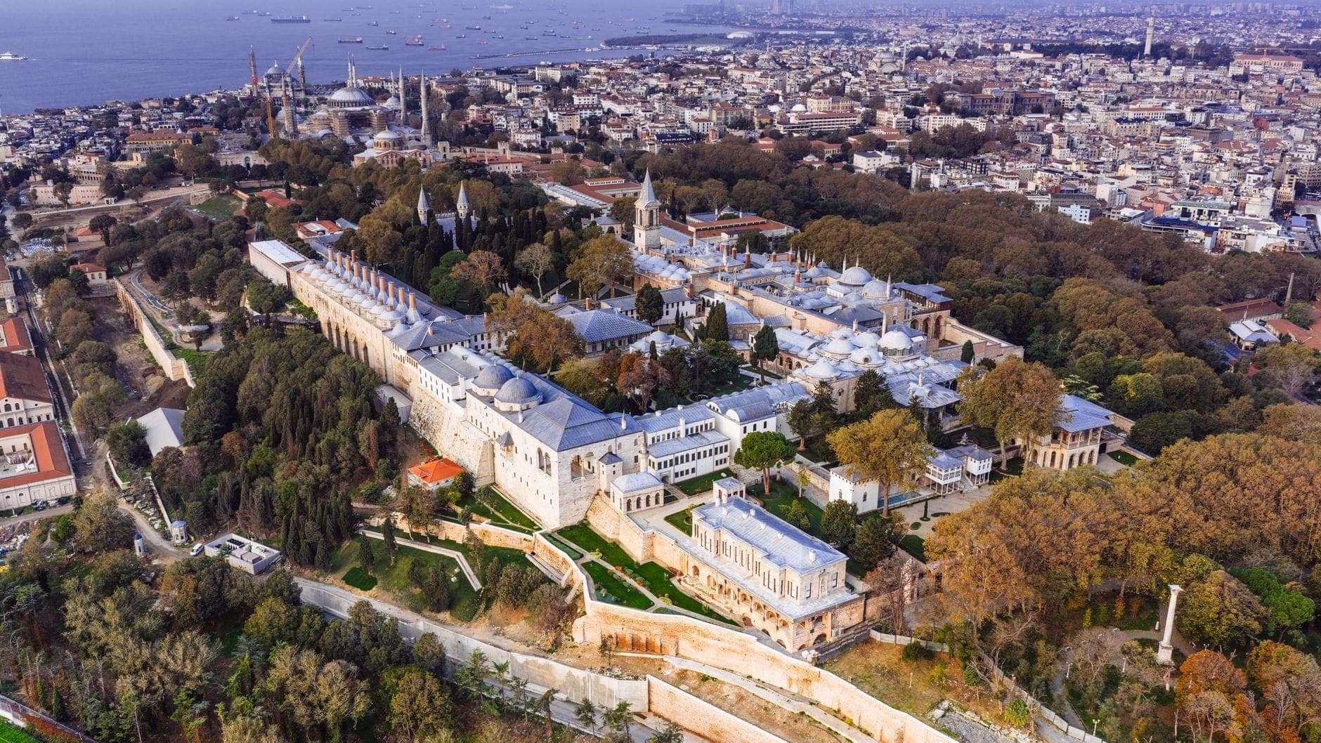 Beeindruckende Luftaufnahme der historischen Halbinsel Istanbuls mit dem weitläufigen Topkapı-Palast inmitten von Bäumen, dahinter der Bosporus und die Stadtsilhouette.