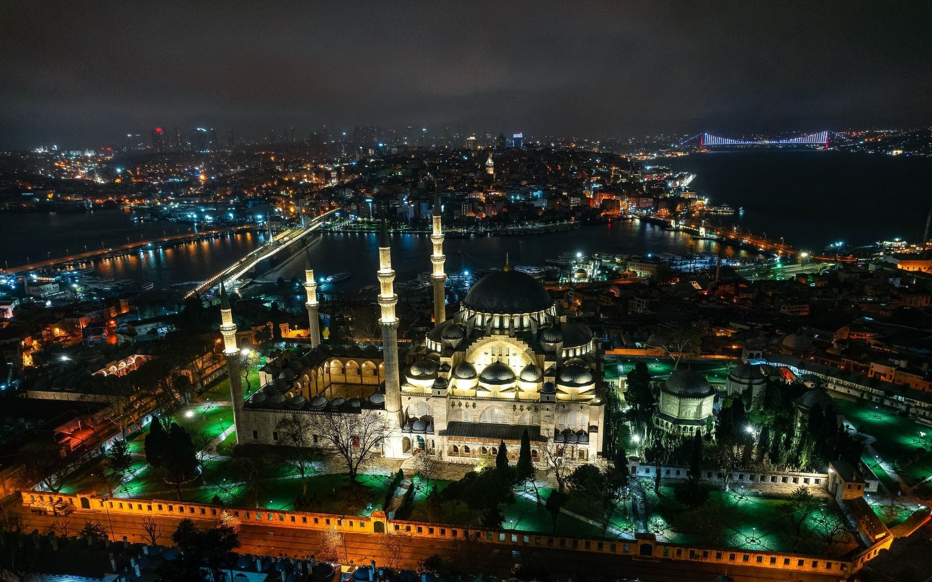 Beeindruckende nächtliche Luftaufnahme einer prachtvoll beleuchteten Moschee in Istanbul, mit der Skyline der Stadt und der Bosporus-Brücke im Hintergrund.