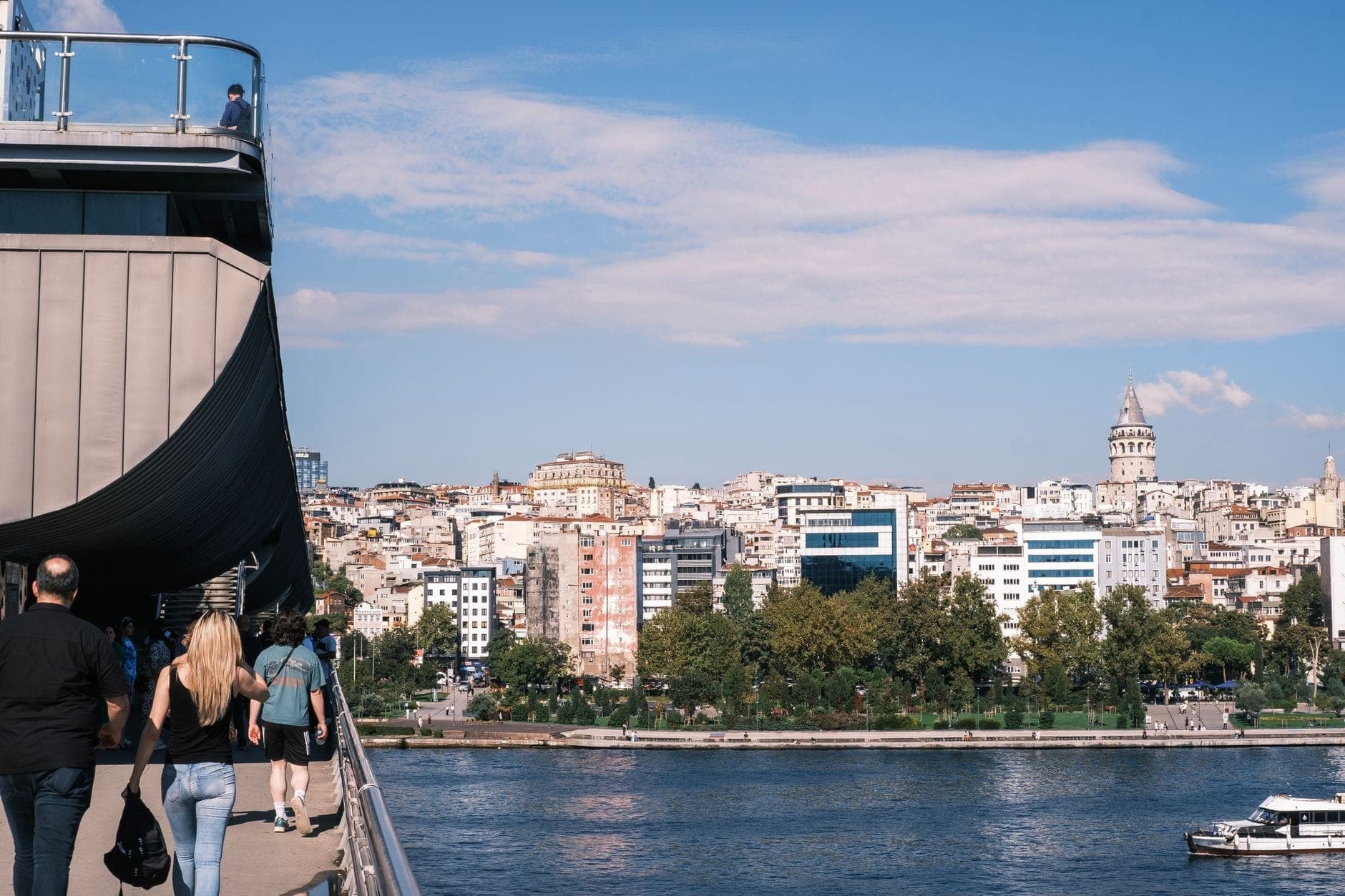 Menschen gehen über eine Brücke über dem Wasser, im Hintergrund die Skyline Istanbuls mit dem Galataturm an einem sonnigen Tag.
