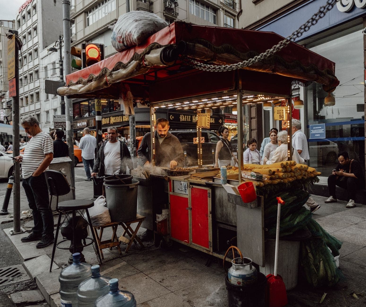 Ein belebter Streetfood-Stand in Istanbul, an dem ein Verkäufer Mais und Kastanien grillt, umgeben von Einheimischen und Besuchern auf einer belebten Stadtstraße.