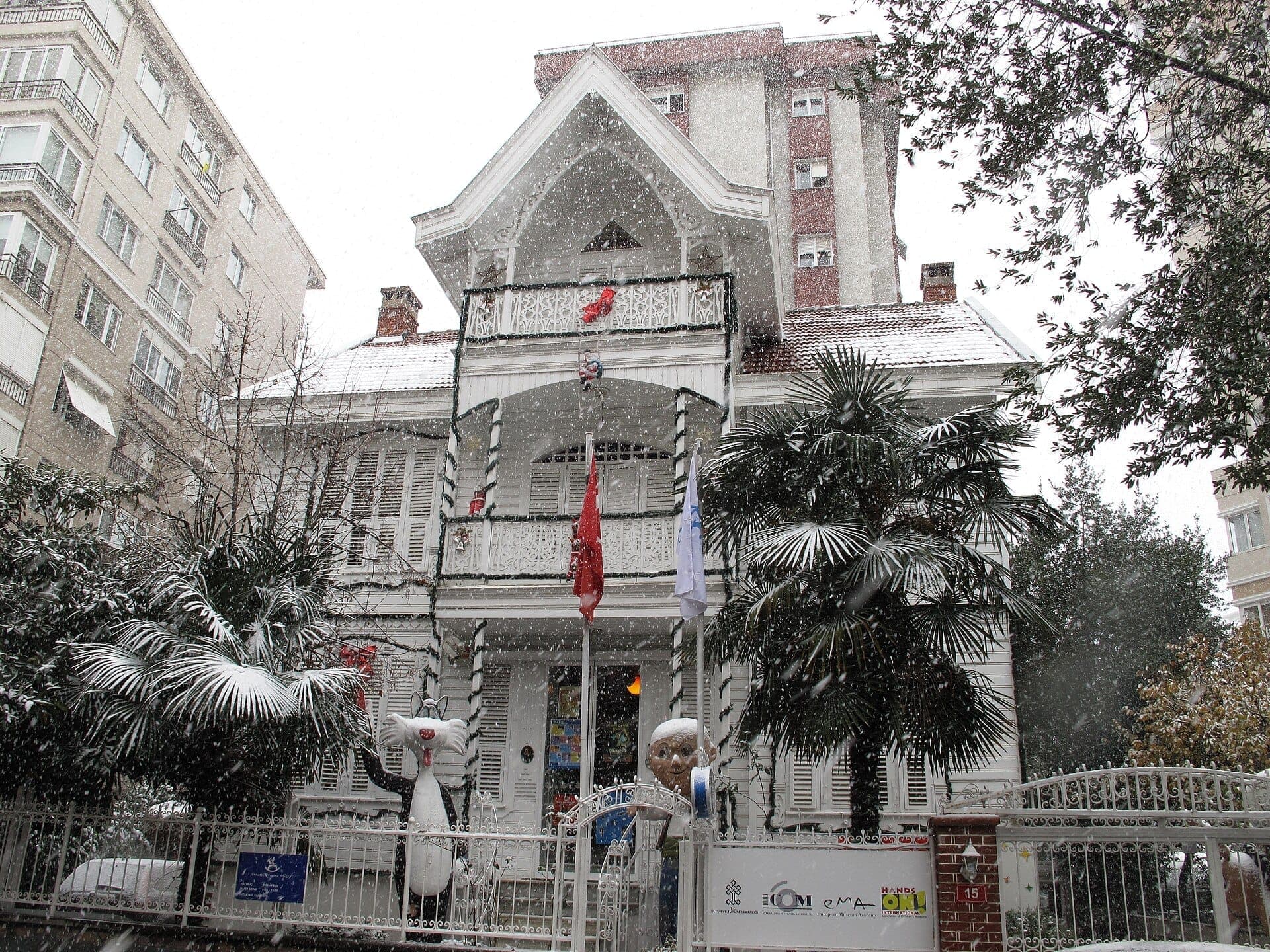 Historisches Holzherrenhaus, in dem sich das Istanbul Toy Museum in Kadıköy befindet, umgeben von verschneiten Palmen und verspielten Spielzeugstatuen am Eingang.