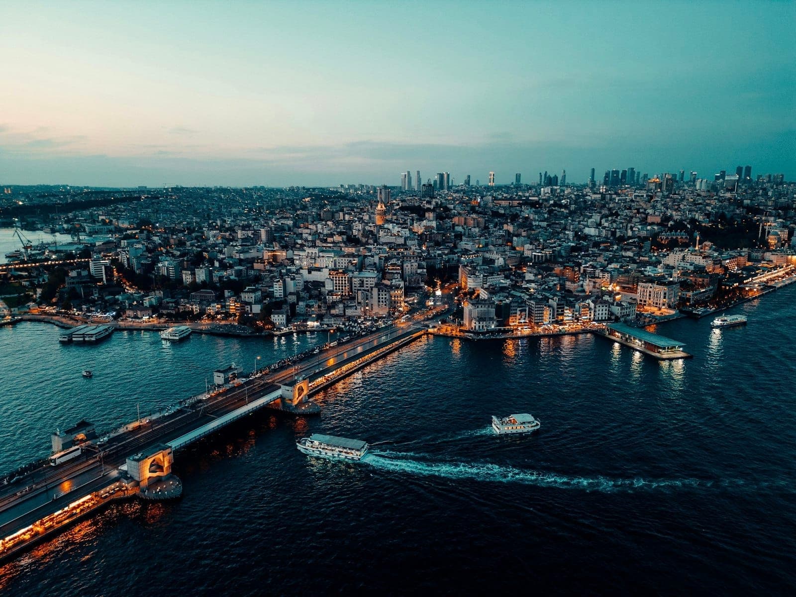Luftaufnahme von Istanbul in der Abenddämmerung mit der beleuchteten Galata-Brücke, Schiffen auf dem Wasser und der Stadtsilhouette mit Moscheen und Dächern.