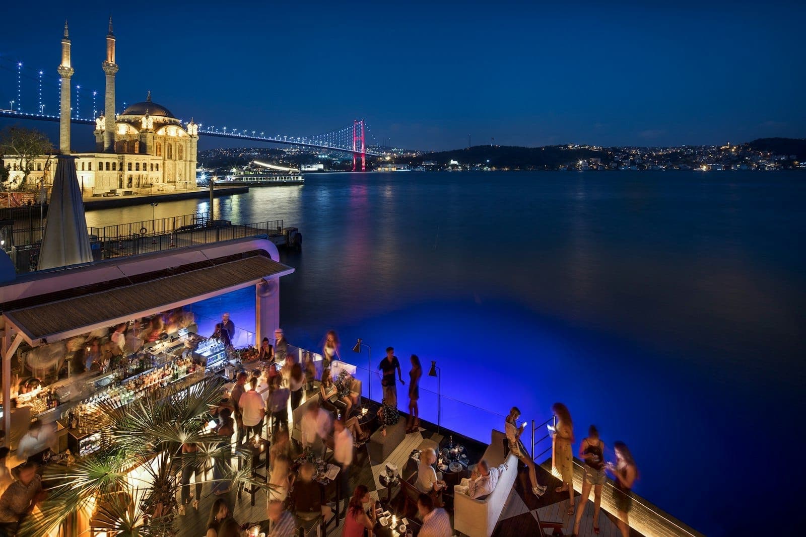 Belebte Dachbar mit Blick auf den Bosporus, lebhaftem Nachtleben, beleuchteter Ortaköy-Moschee und Bosporus-Brücke am Abend, Istanbul.