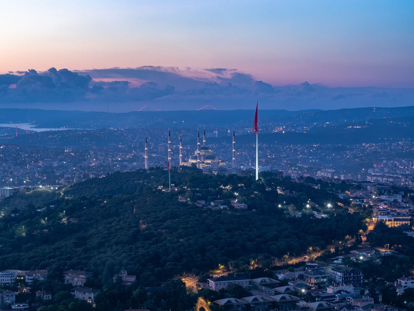 Panoramablick auf Istanbul in der Abenddämmerung, mit einem bewaldeten Hügel mit einer großen Moschee, türkischer Flagge und der Skyline im Hintergrund.