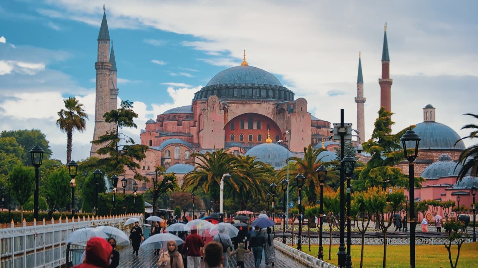 Blick auf die Hagia Sophia mit Menschen, die auf der Hauptpromenade spazieren, einige mit Regenschirmen, üppiger Bepflanzung und dramatischem Himmel in Sultanahmet.