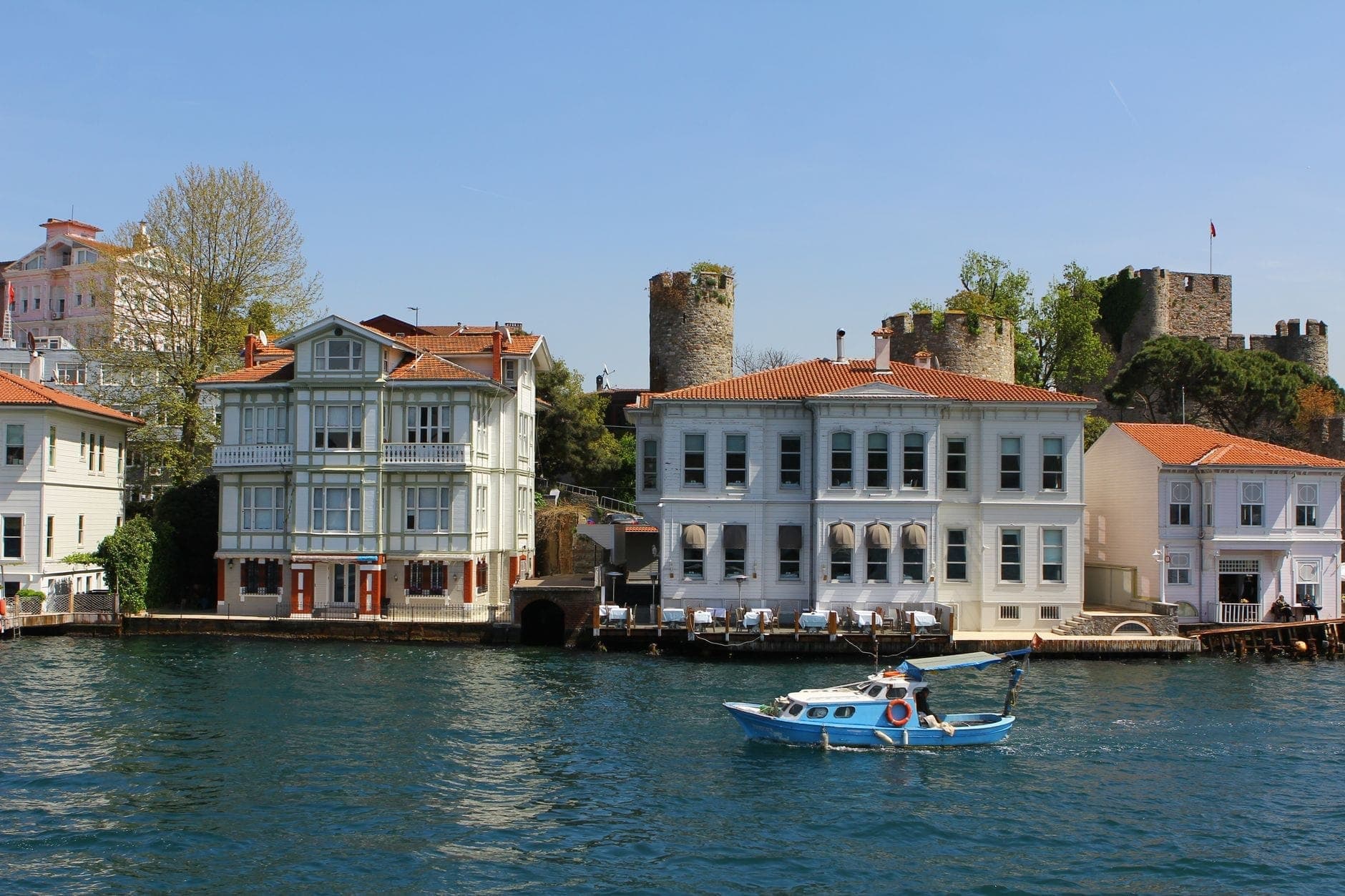 Hölzerne Yalı-Häuser und Festungsruinen entlang der Uferpromenade des Bosporus in Istanbul an einem klaren Tag, mit einem kleinen Boot davor.