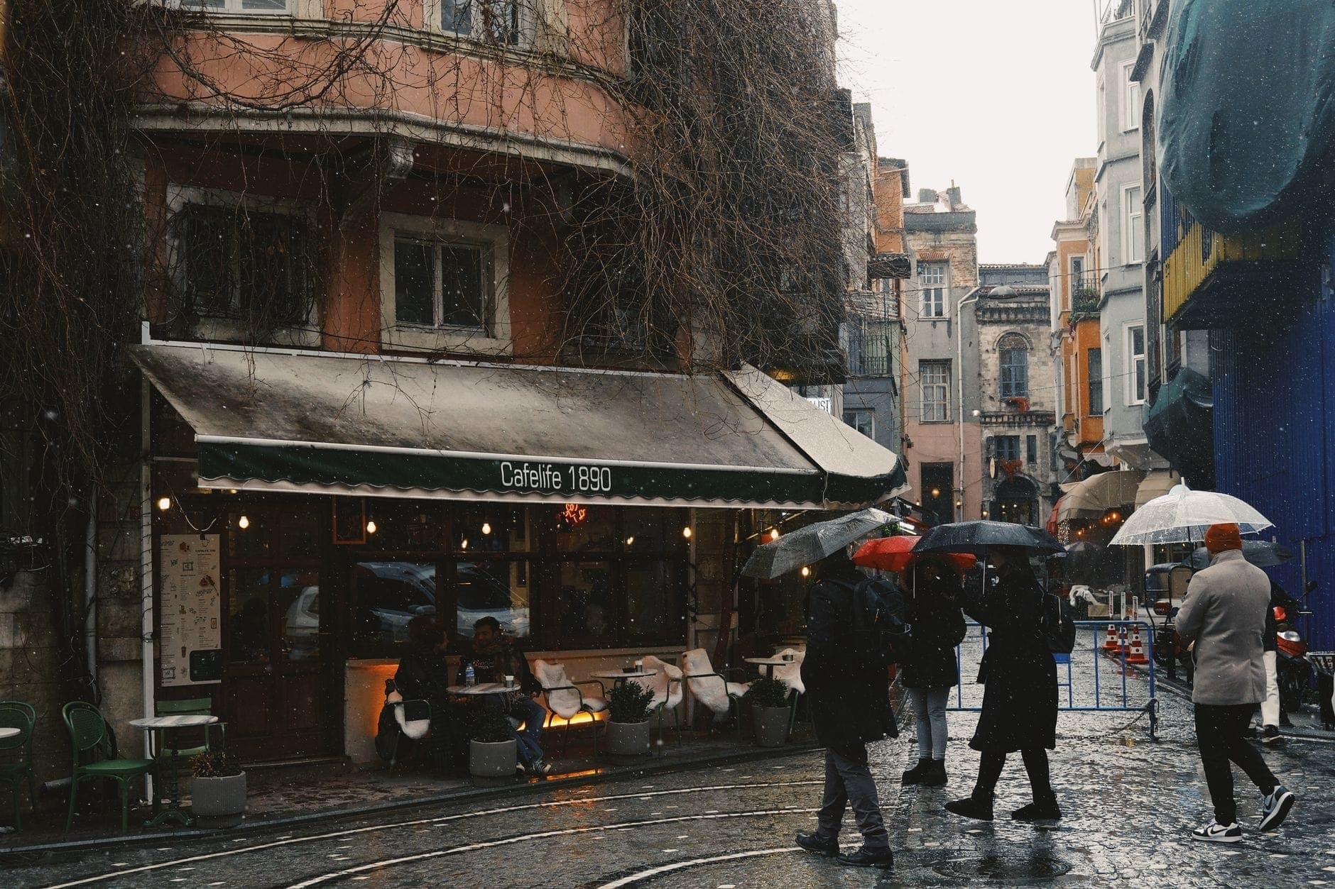 Personas caminando con paraguas por una calle mojada en Estambul cerca de un café, mostrando el clima lluvioso y la vida urbana.