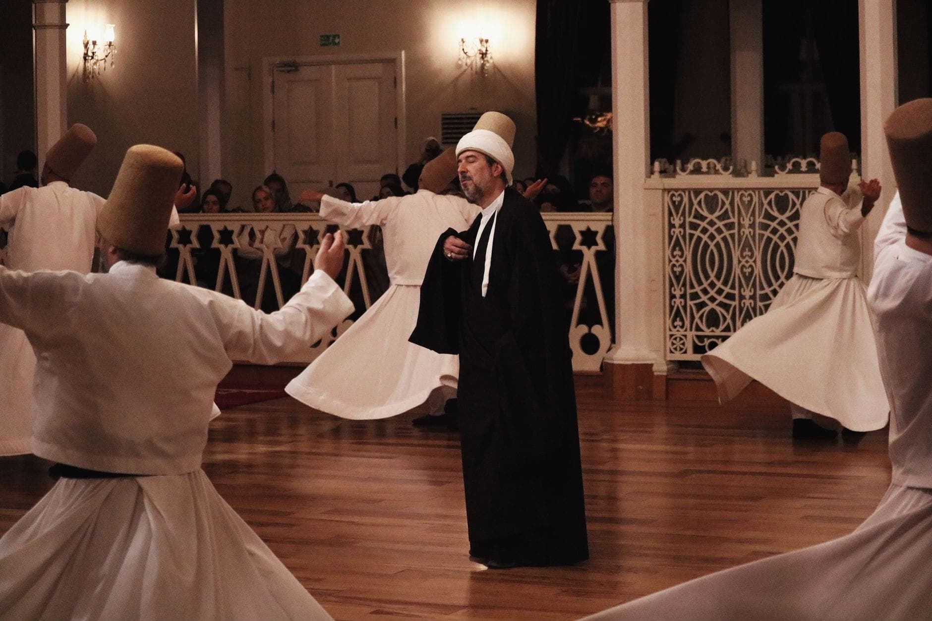 Tanzende Derwische bei der Sema-Zeremonie in einem eleganten Saal, in traditionellen weißen Gewändern und hohen Hüten, mit Zuschauern im Hintergrund.