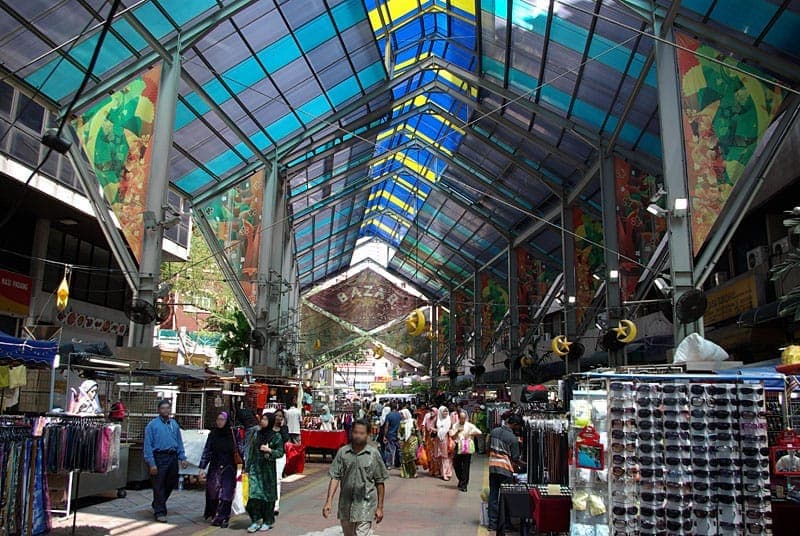 Cảnh chợ có mái che trên Jalan Masjid India với mái kính đầy màu sắc, các gian hàng nhộn nhịp bán vải và kính mát, và người đi bộ dọc theo hành lang.