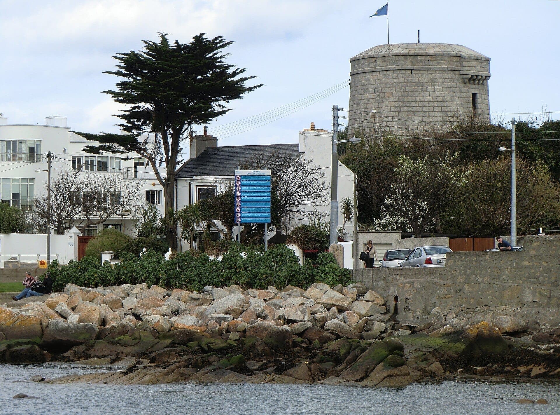 Der James Joyce Tower & Museum, ein runder steinerner Martello-Turm, steht an felsigen Küstenlinien, mit modernen Häusern, Grünflächen und einem blauen Informationsschild in der Nähe.