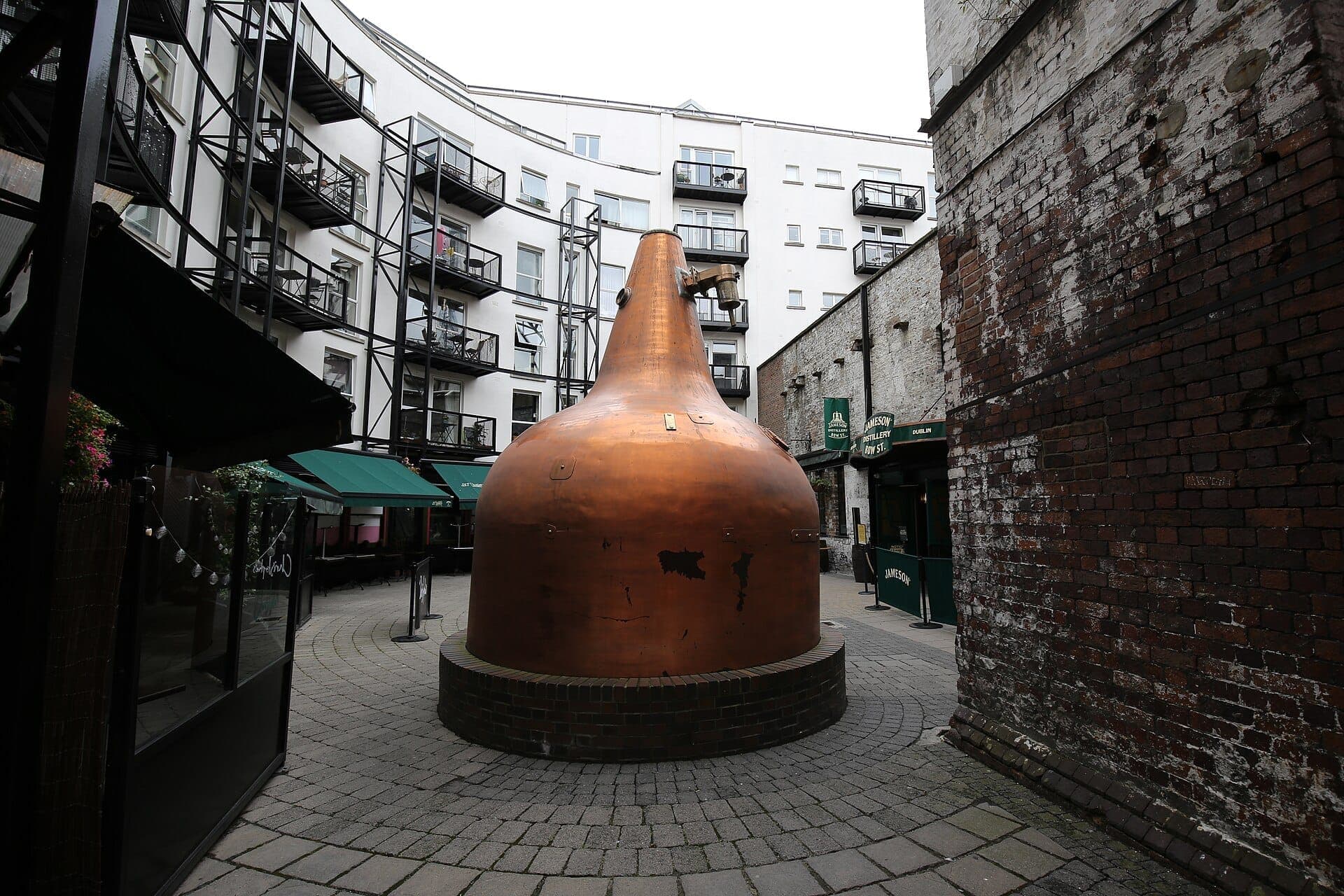 Duży miedziany alembik przed destylarnią Jameson Bow St, otoczony historyczną ceglaną zabudową i nowoczesnymi apartamentami w dzielnicy Smithfield w Dublinie.