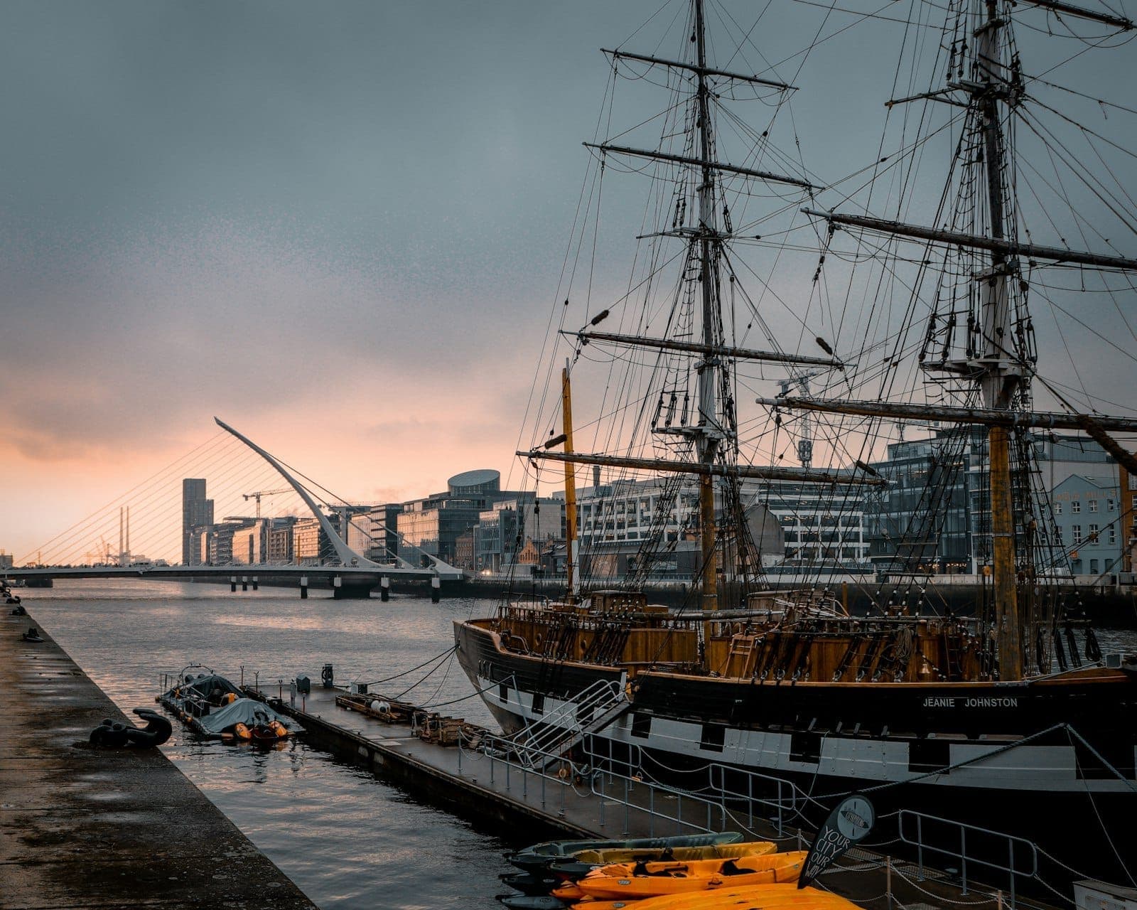 Żaglowiec Jeanie Johnston zacumowany w dublińskich Docklands o zachodzie słońca, z panoramą miasta i mostem Samuela Becketta w tle.