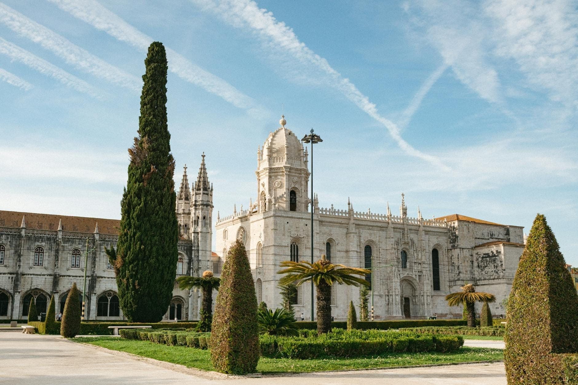 Toàn cảnh tu viện Jerónimos ở Lisbon với kiến trúc Manueline tinh xảo, tháp cao, vườn cây xanh và bầu trời xanh điểm vài đám mây nhẹ cùng vệt khói máy bay.
