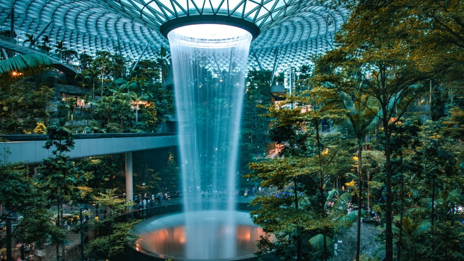 Wide landscape view of Jewel Changi Airport’s Rain Vortex waterfall cascading through lush indoor forest beneath the glass dome, with skytrain and visitors visible among greenery.