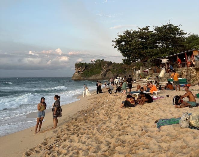 Mga bisita na nagpapahinga sa puting buhangin ng Balangan Beach sa Jimbaran, Bali, na naka-frame ng mga dramatikong limestone cliff, beachside warung, at mahinahong alon sa ilalim ng malinaw na langit