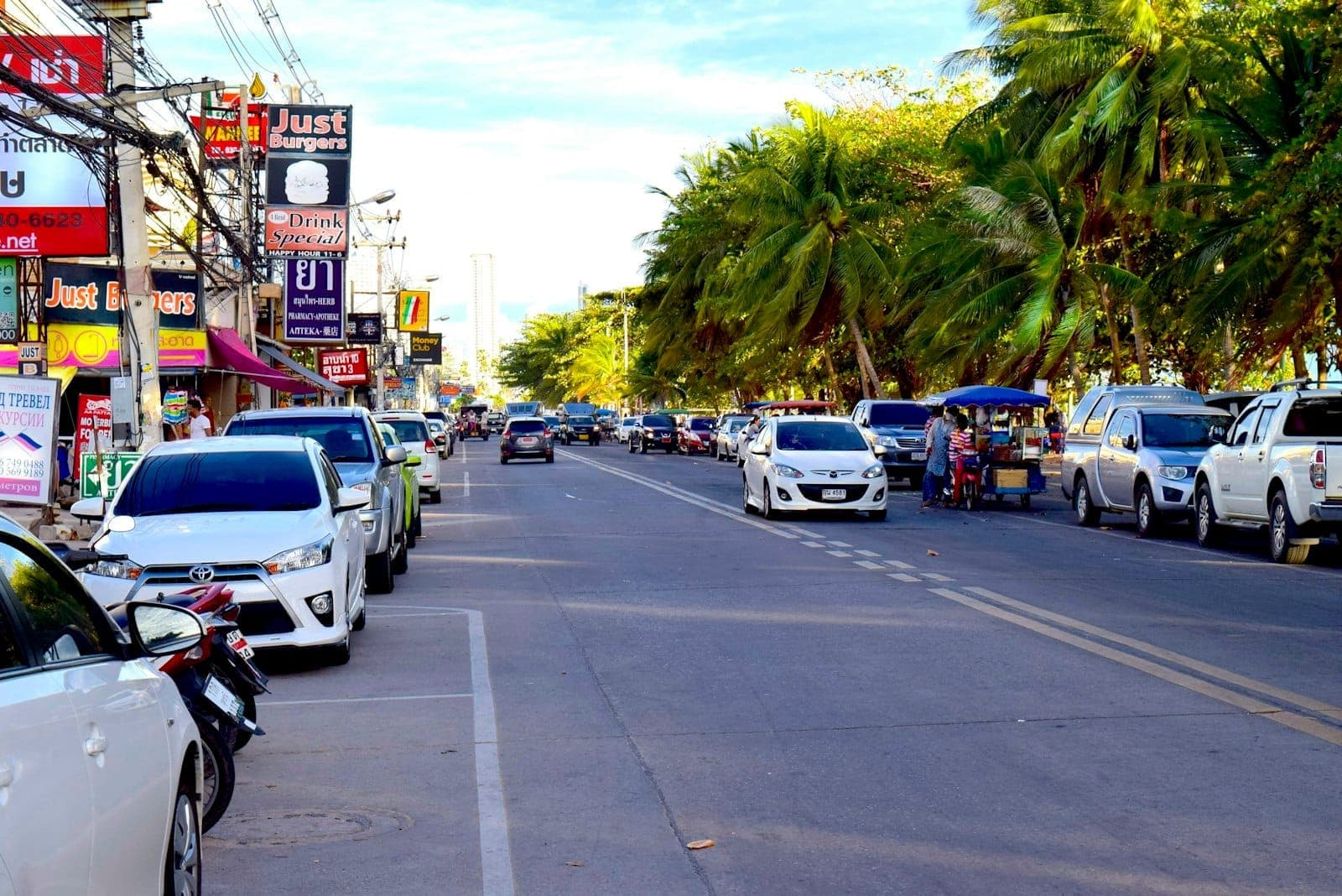 Busy street in Jomtien, Pattaya lined with parked cars, local shops, restaurants, and tall palm trees under a bright afternoon sky.