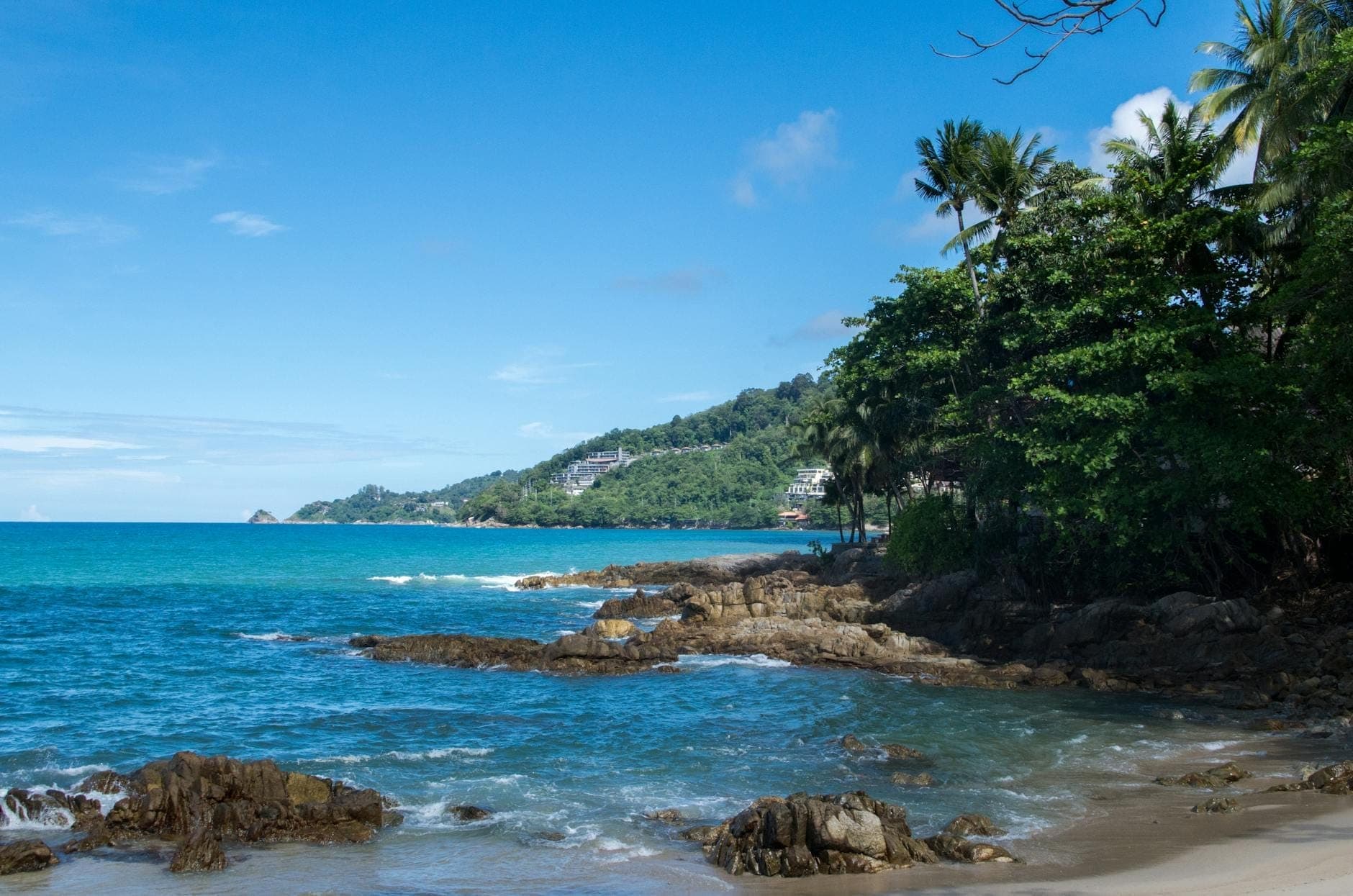 Tanawin ng isang tahimik na dalampasigan sa Phuket na may turquoise na tubig, batuhan, malulusog na burol na may kagubatan, at mga puno ng palma sa ilalim ng maliwanag na asul na langit na may kalat-kalat na ulap.
