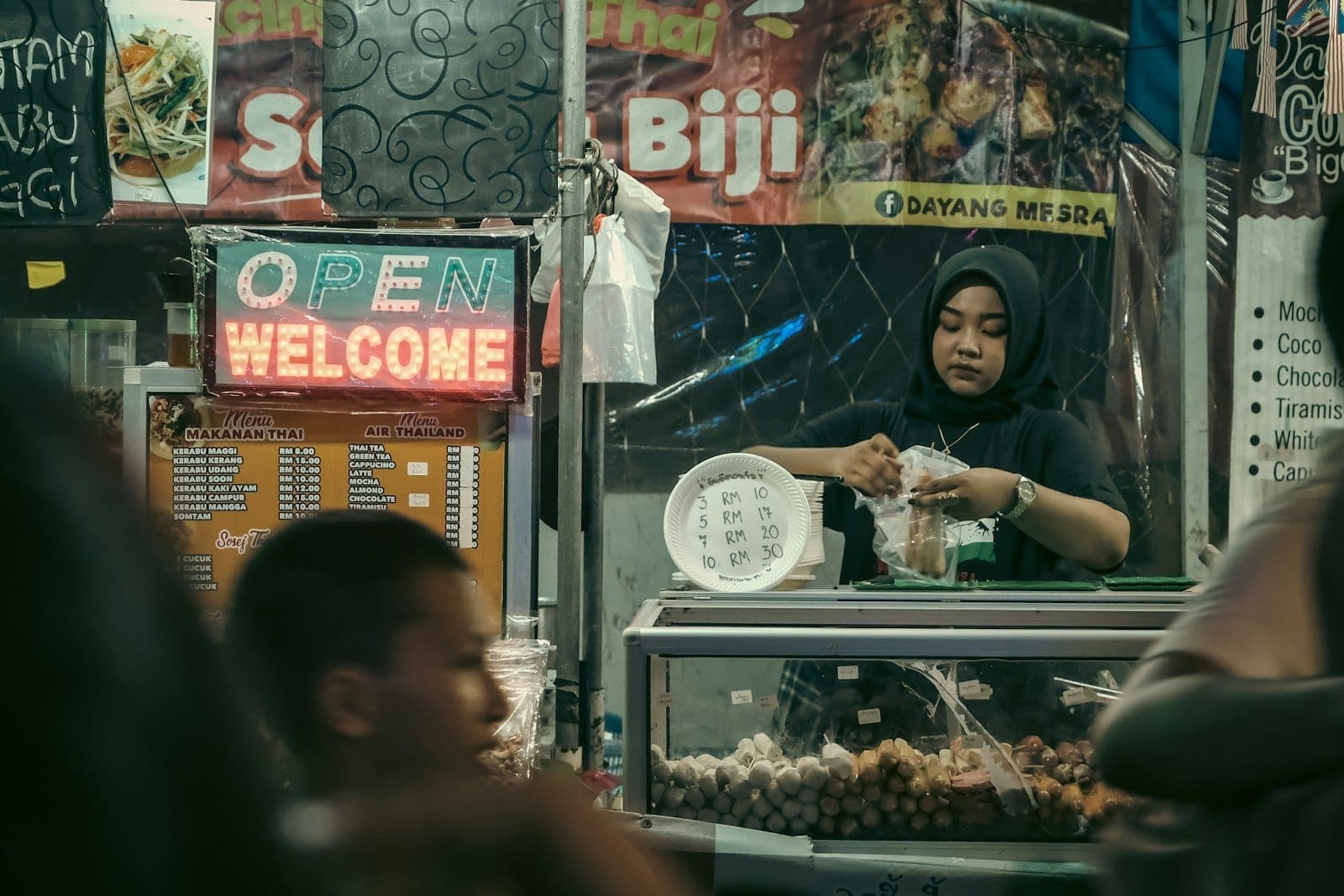 Góc nhìn của du khách tại chợ đường phố Kampung Baru, nổi bật với hình ảnh người bán hàng đóng gói thức ăn trong khi du khách khám phá các món đặc sản Mã Lai địa phương.